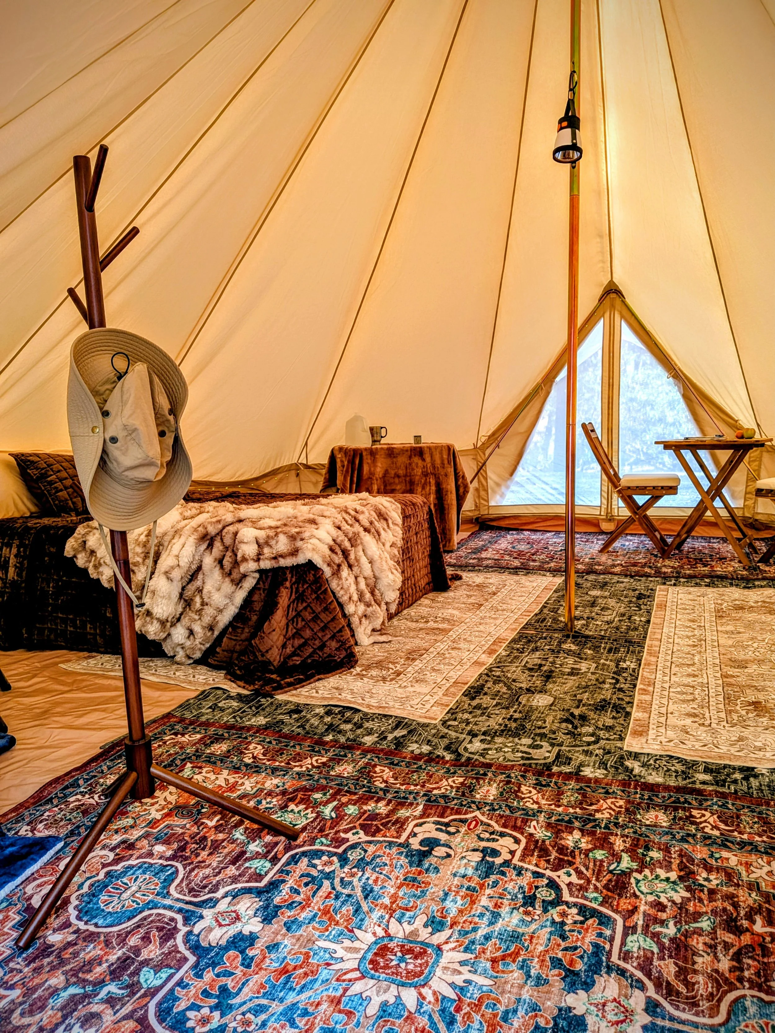 Interior of a cozy glamping bell tent with patterned rugs, a bed with a faux fur throw, a table with cups, and a small dining area with two chairs.