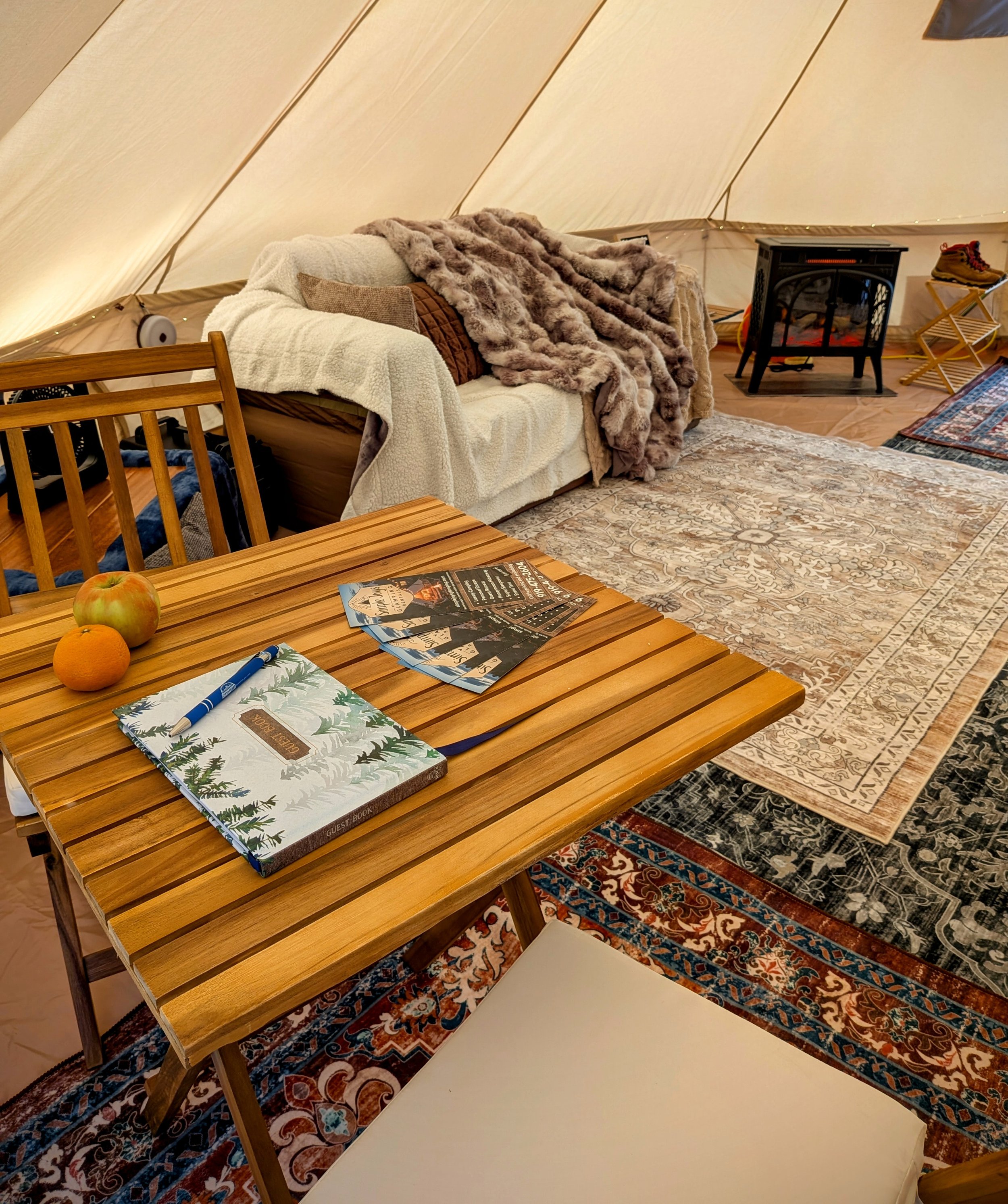 Cozy indoor living space with a wooden table, a guest book, magazine pamphlets, and a white pen. In the background, a white couch is draped with blankets and pillows, with plush throws on top, under a tent-like ceiling. The floor has patterned rugs, 