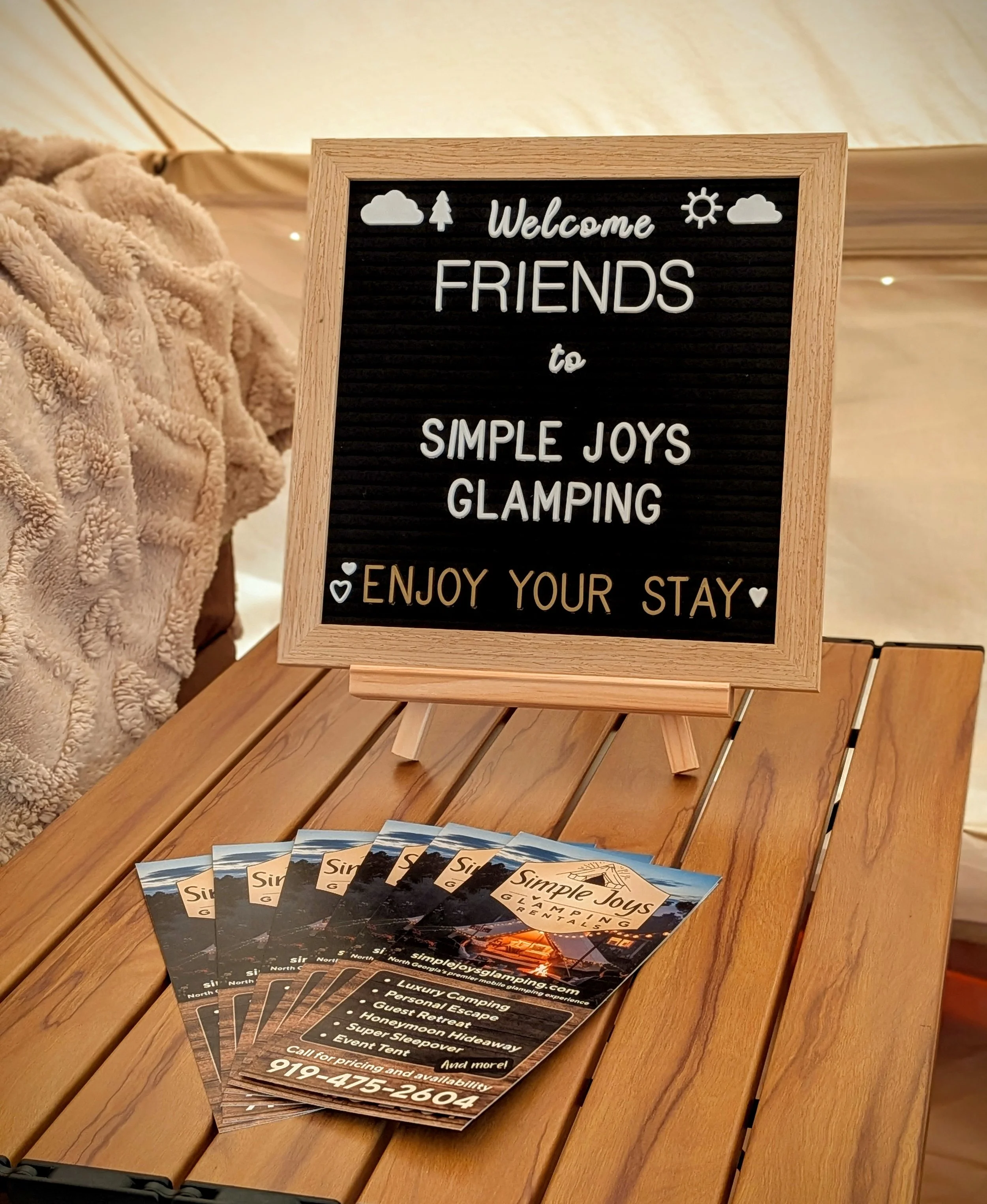 Signboard welcoming friends to Simple Joys Glamping and promotional flyers for Simple Joys Glamping with camping amenities, placed on a wooden table, inside a cozy bell tent.