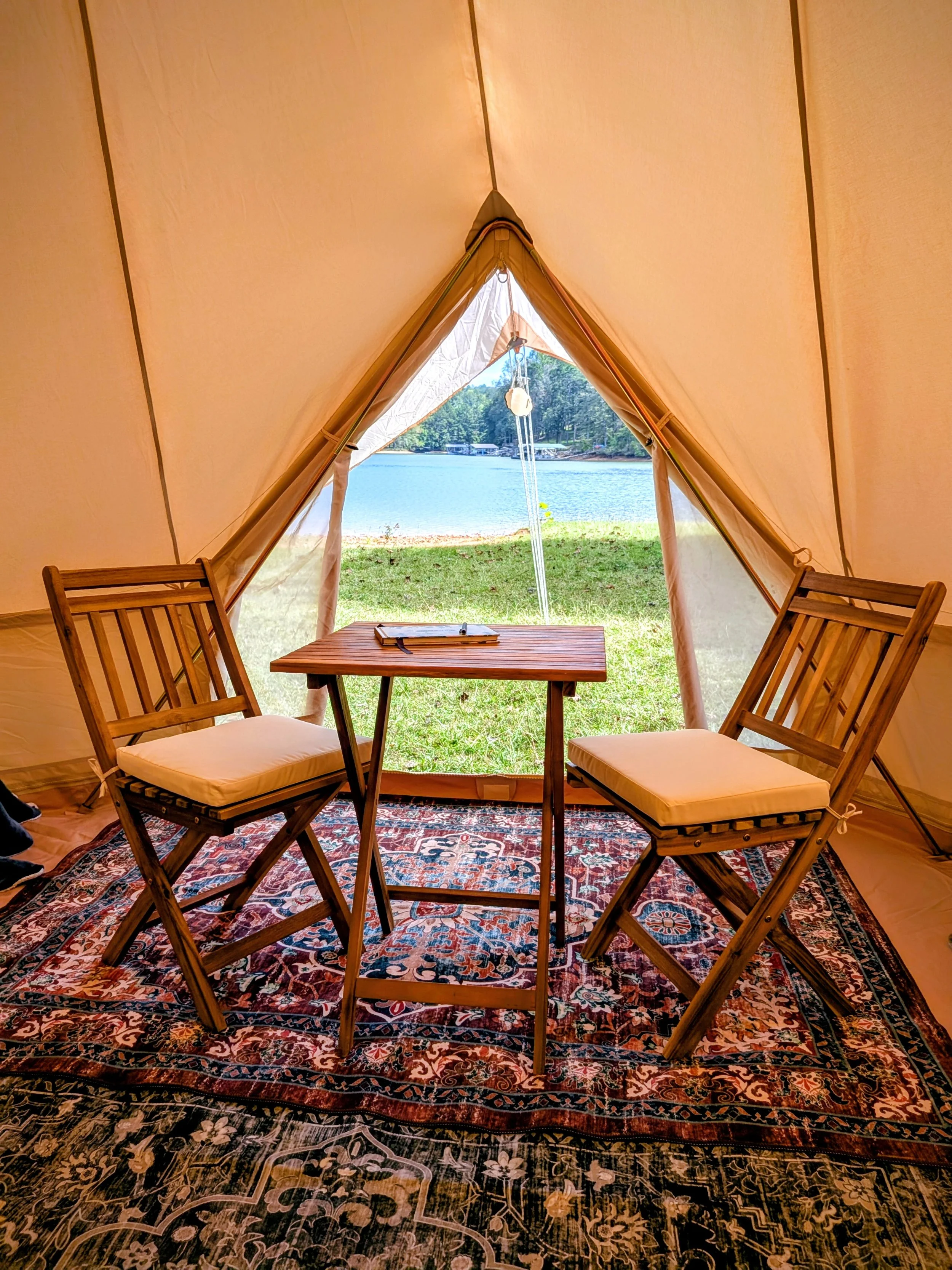 Inside a beige glamping bell tent facing a lake with two wooden chairs and a small wooden table. There is a rug underneath the table and chairs, and a guest book with a pen on the table.