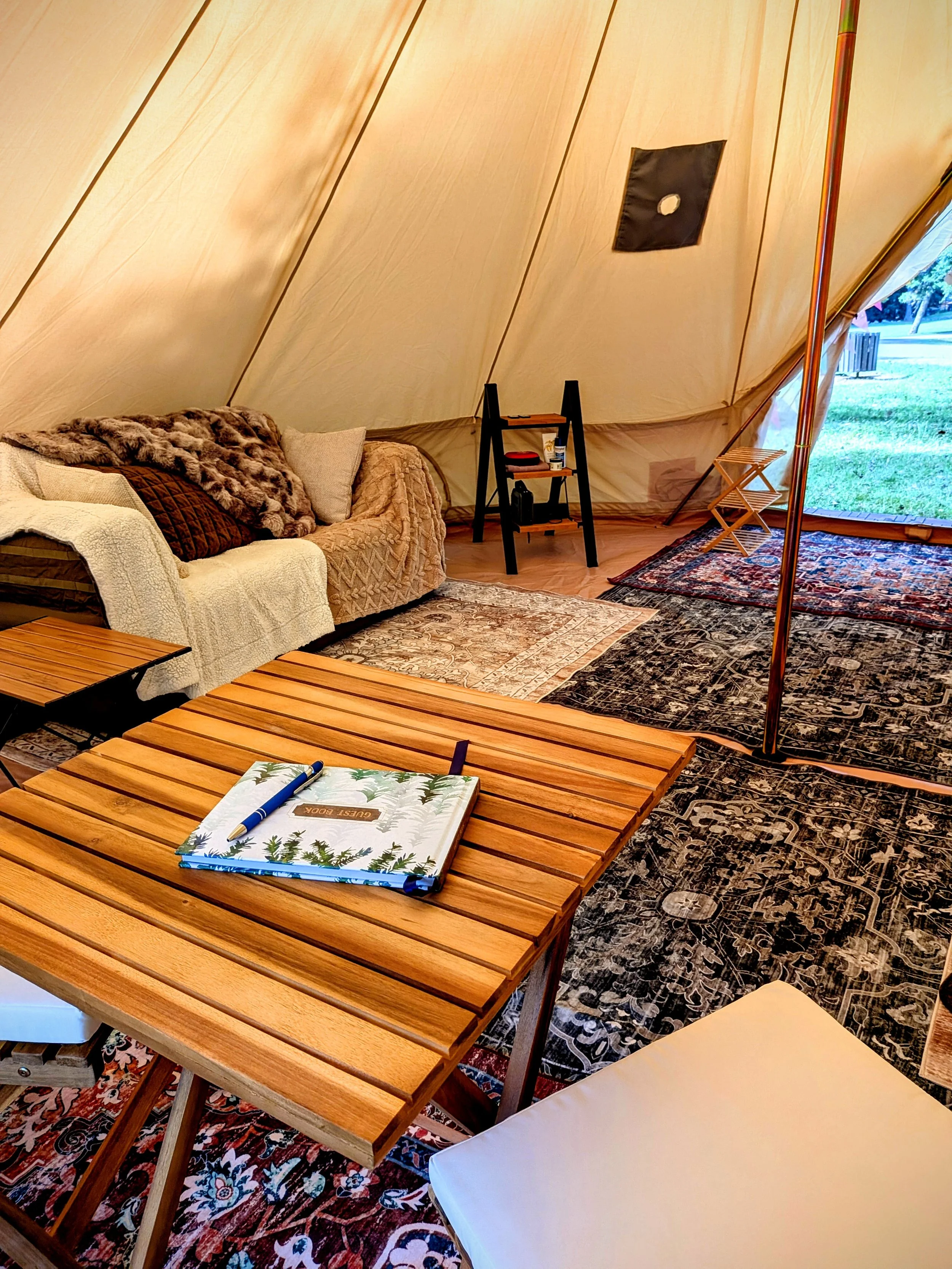 Interior of a cozy glamping bell tent with a wooden table, a sofa with cushions and blankets, patterned rugs, a bookshelf, and an open flap showing a grassy outdoor area.
