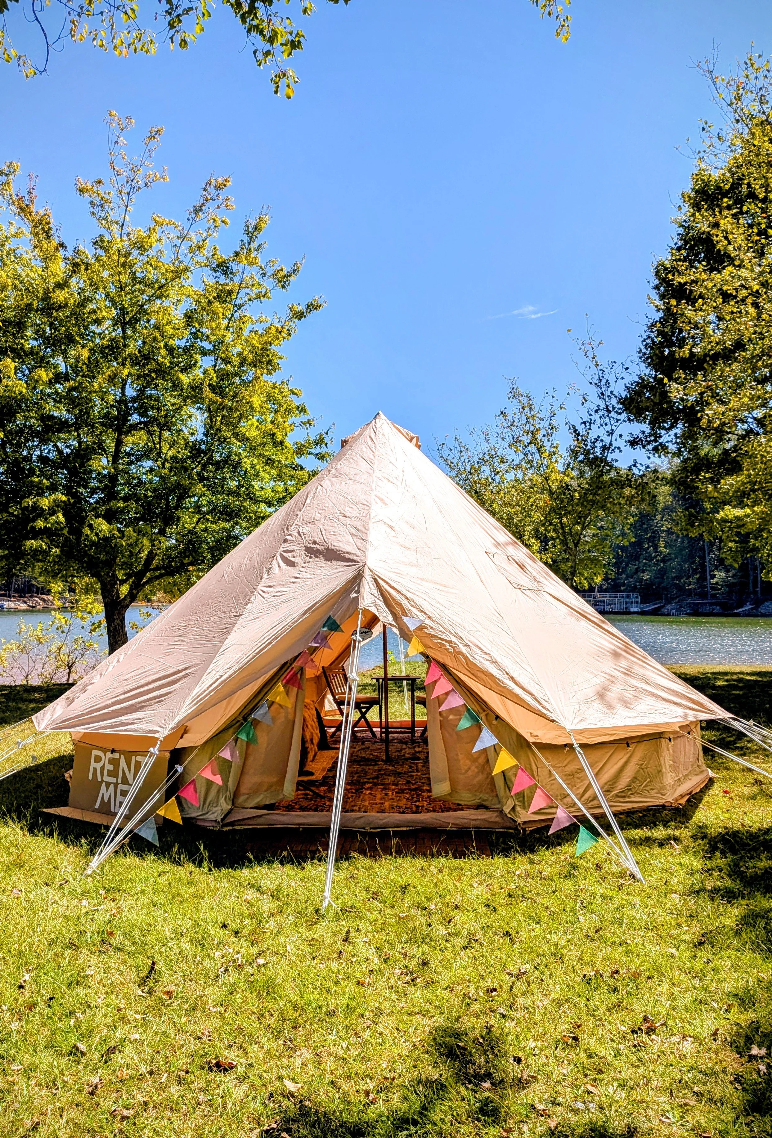 A beige canvas glamping bell tent set up on a grassy area by a lake, decorated with colorful pennant banners, with trees and a lake in the background and a clear blue sky.