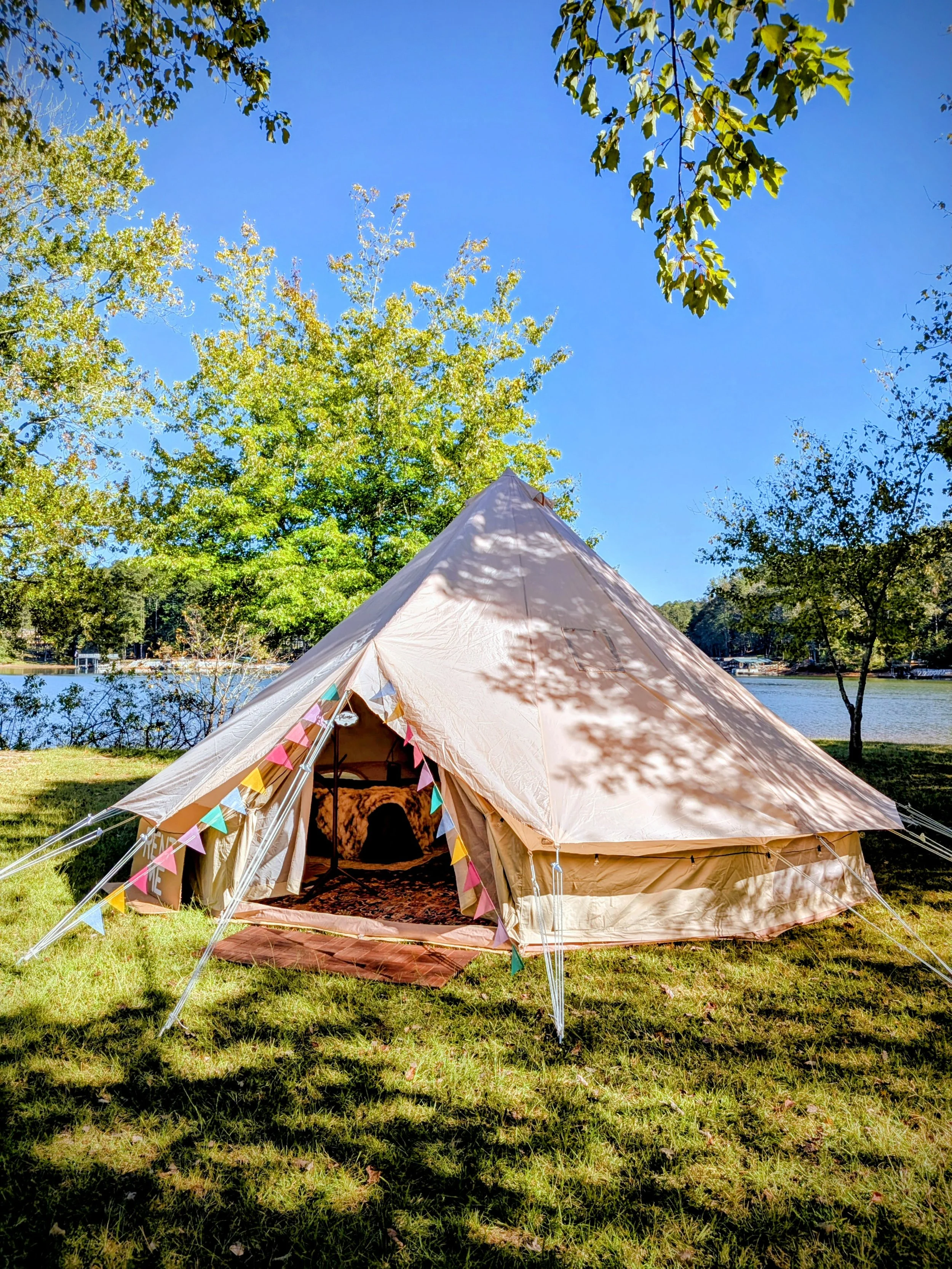 A beige bell tent set up on grass near a lake with trees and blue sky overhead, decorated with colorful bunting.