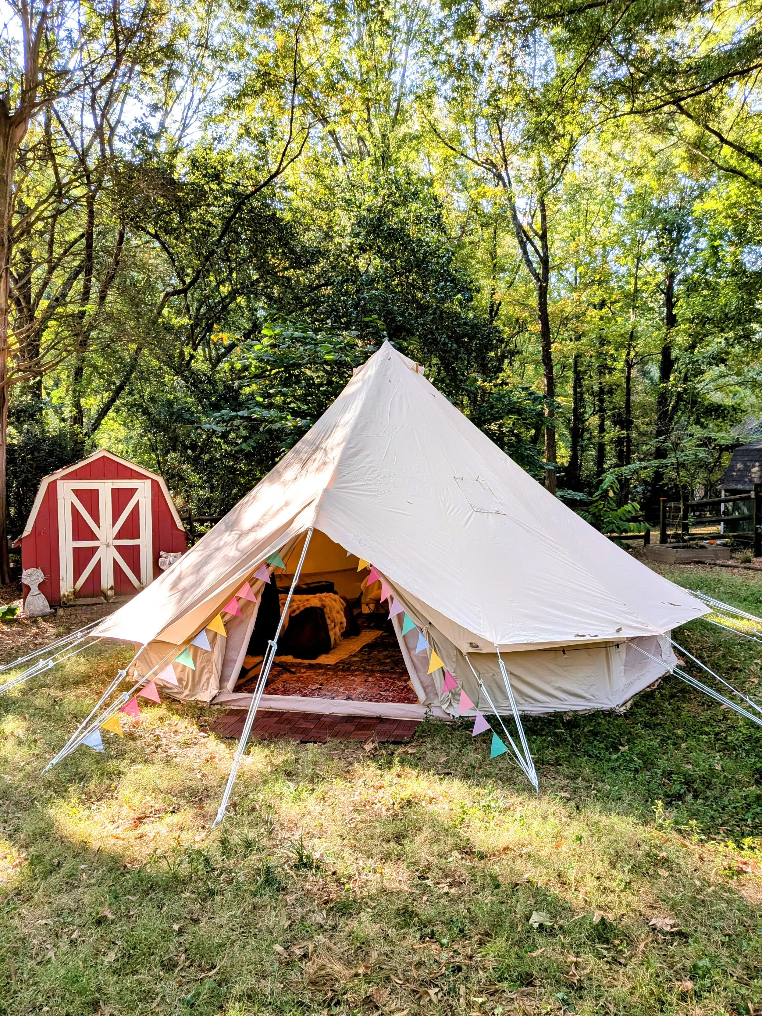 A beige glamping bell tent decorated with colorful pennants on a grassy area, with a red shed and green trees in the background.