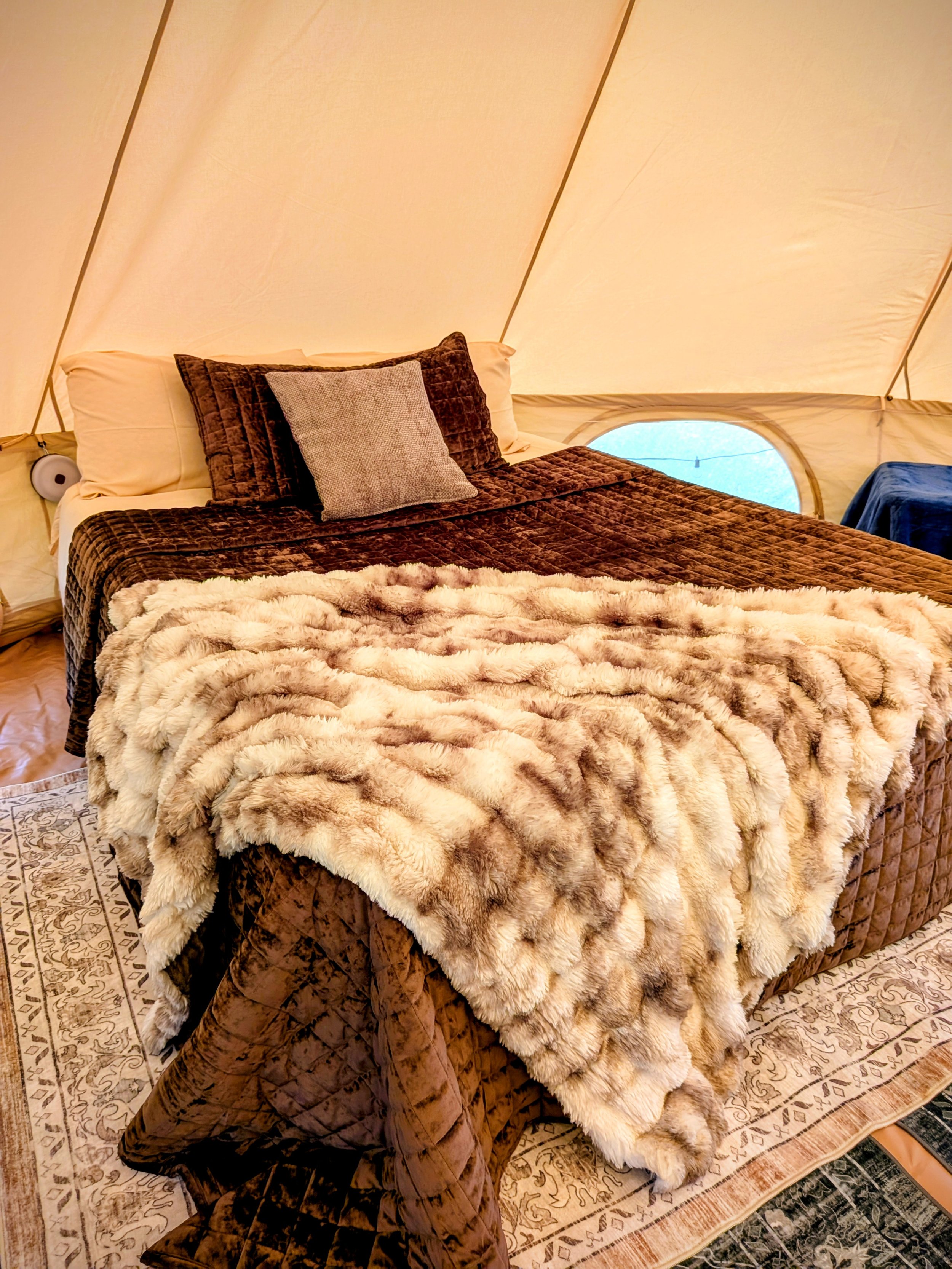 A cozy bed inside a glamping bell tent, with brown velvet bedding, pillows, and a plush cream-colored blanket.