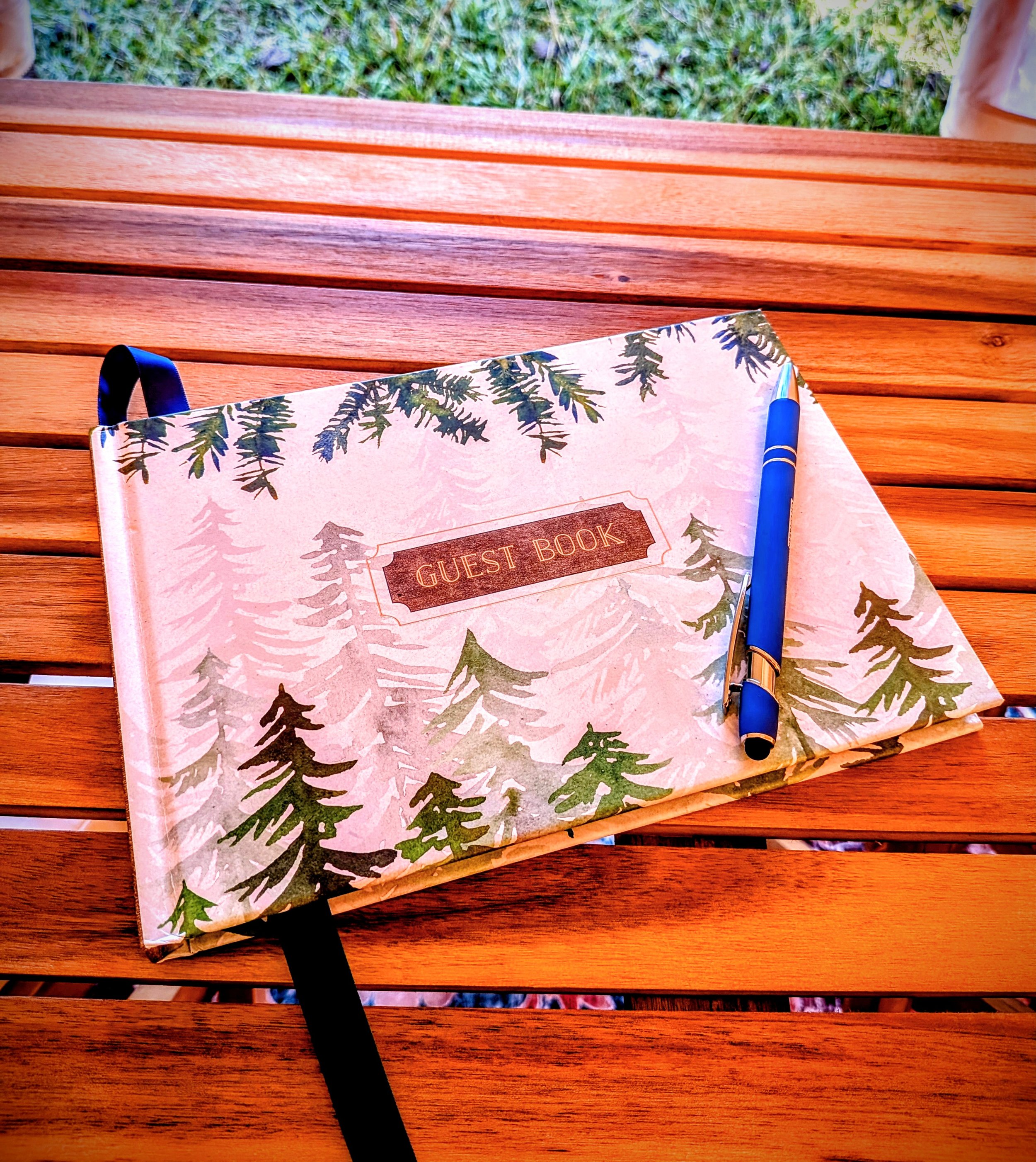 Guest book with a forest tree design cover and a blue pen resting on it, placed on a wooden outdoor table.