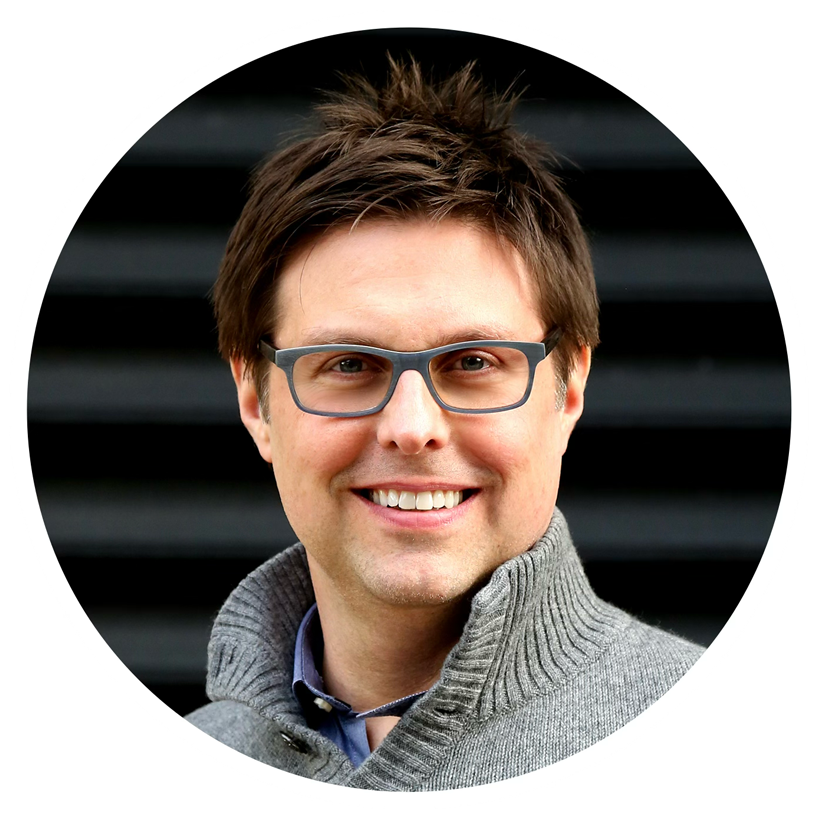 A smiling man with brown hair, wearing glasses and a gray knit sweater, standing outdoors in front of a dark background.