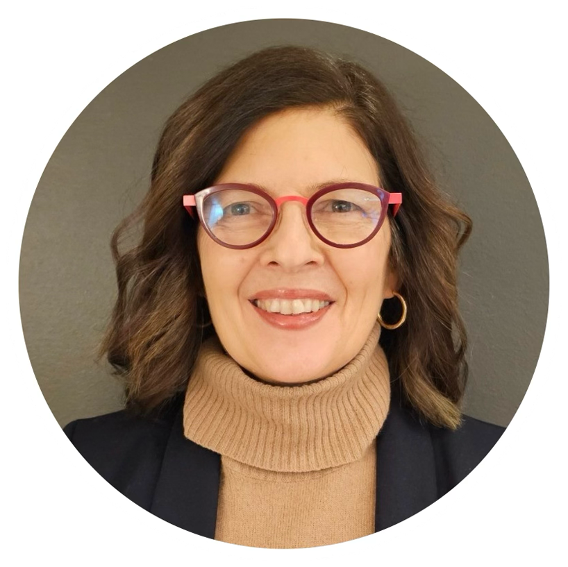 A woman with shoulder-length brown wavy hair and pink glasses, smiling, wearing a tan turtleneck sweater and a black blazer, with gold hoop earrings, standing against a neutral background.