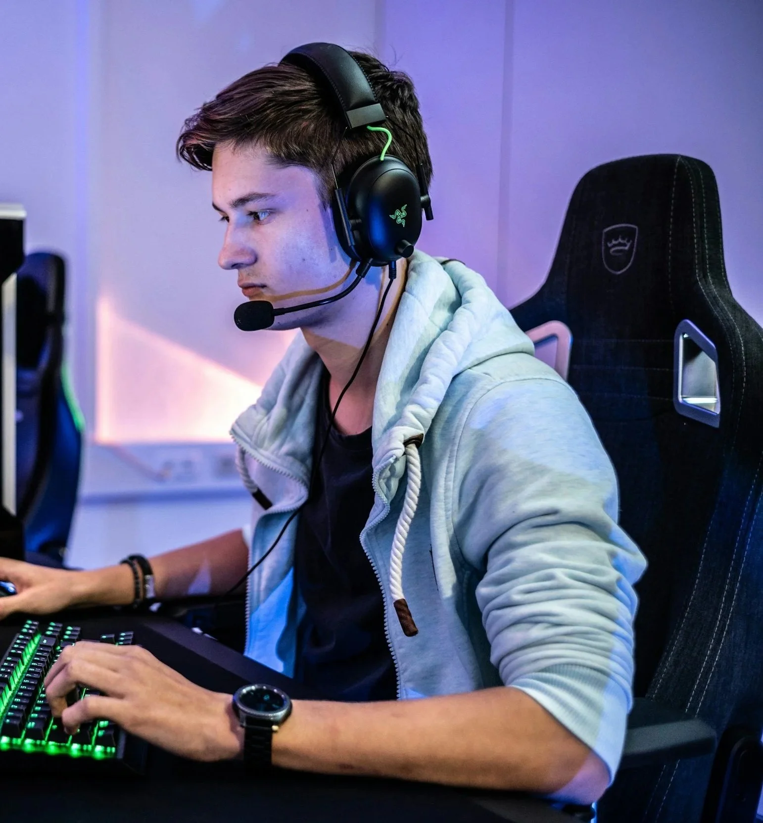 Young man gaming at his computer wearing a headset with microphone, sitting in a gaming chair, using a backlit keyboard.