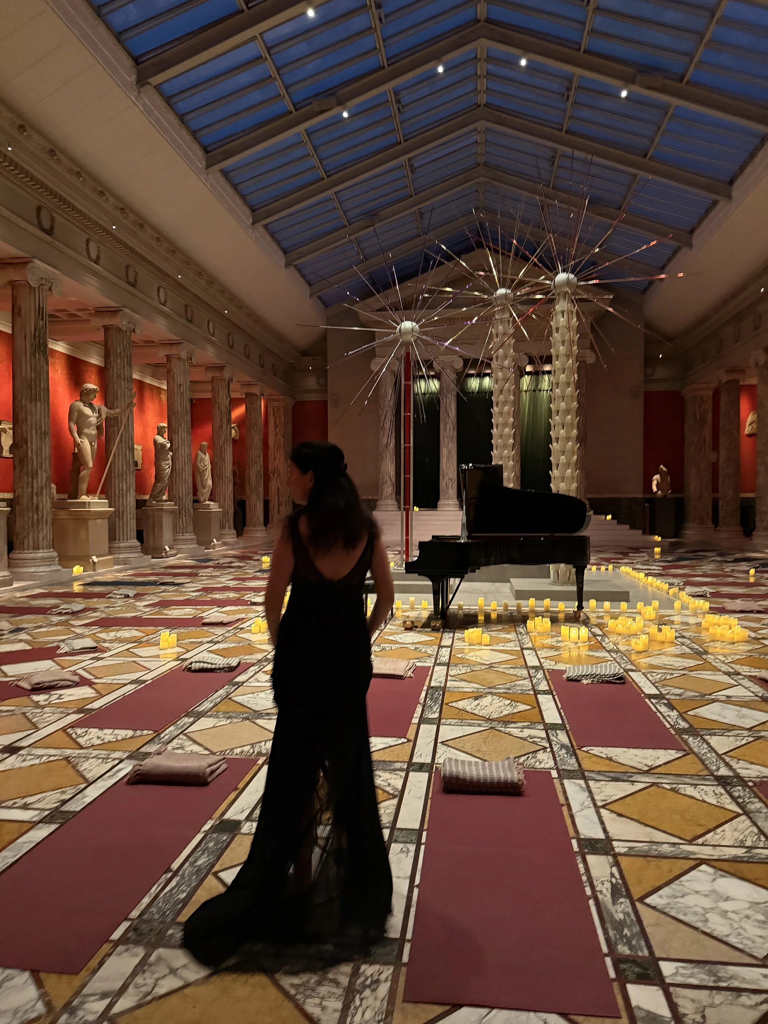 A woman in a black dress stands in a decorated indoor space with a grand piano, surrounded by yoga mats and pillows, with candles, sculptures, columns, and an art installation of large dandelion-like structures overhead.