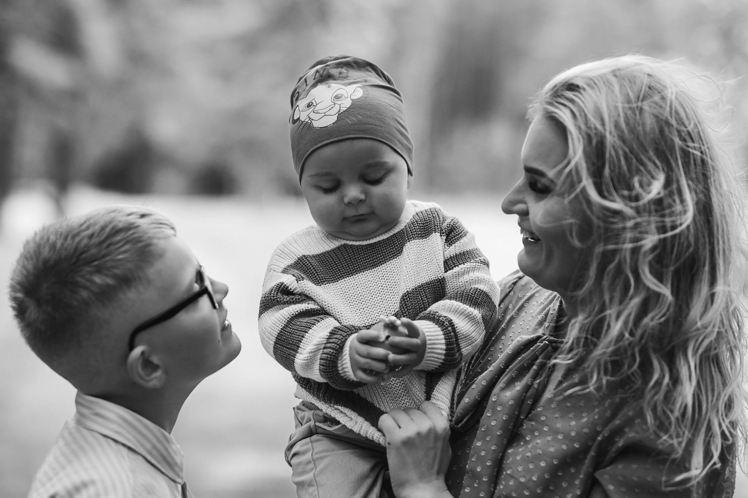 A woman smiling and holding a young child wearing a striped sweater and a beanie hat, while a boy with glasses looks on, in an outdoor setting.