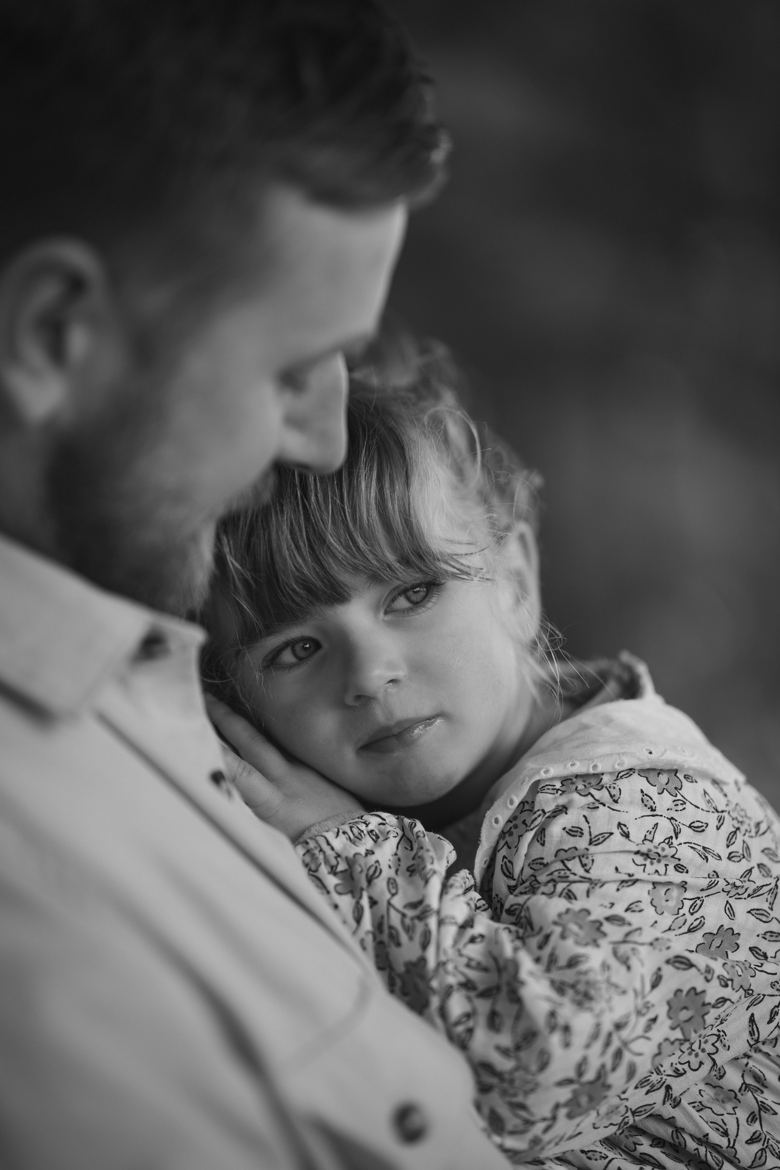 A black and white photo of a bearded man holding a young girl with curly hair and large eyes, resting her head on his shoulder, looking at him with a gentle expression.