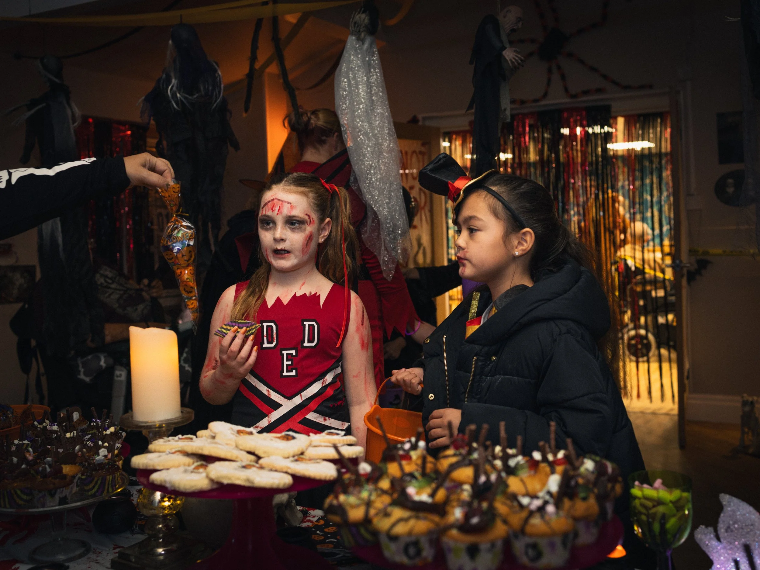 Two young girls dressed in Halloween costumes stand at a table filled with Halloween-themed treats during a Halloween party. One girl has face paint resembling a zombie or monster, while the other girl is wearing a black jacket and headband with a bow. The background features hanging Halloween decorations and a dimly lit room.