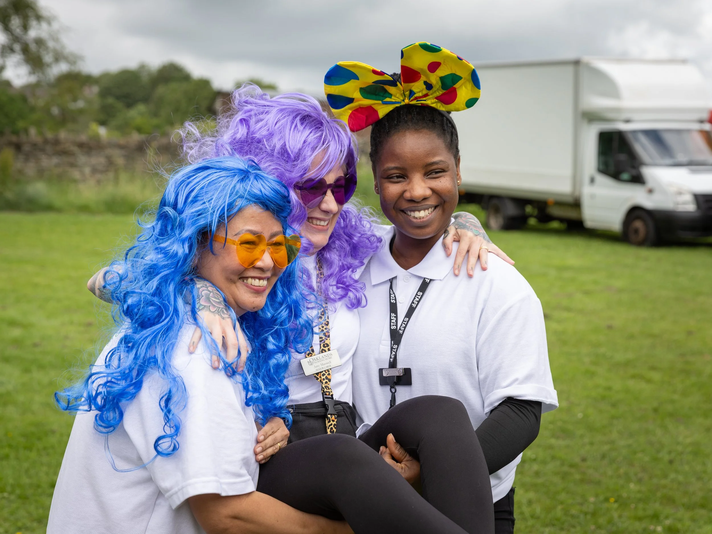 Three women in colorful wigs and glasses smiling and embracing outdoors near a white truck.