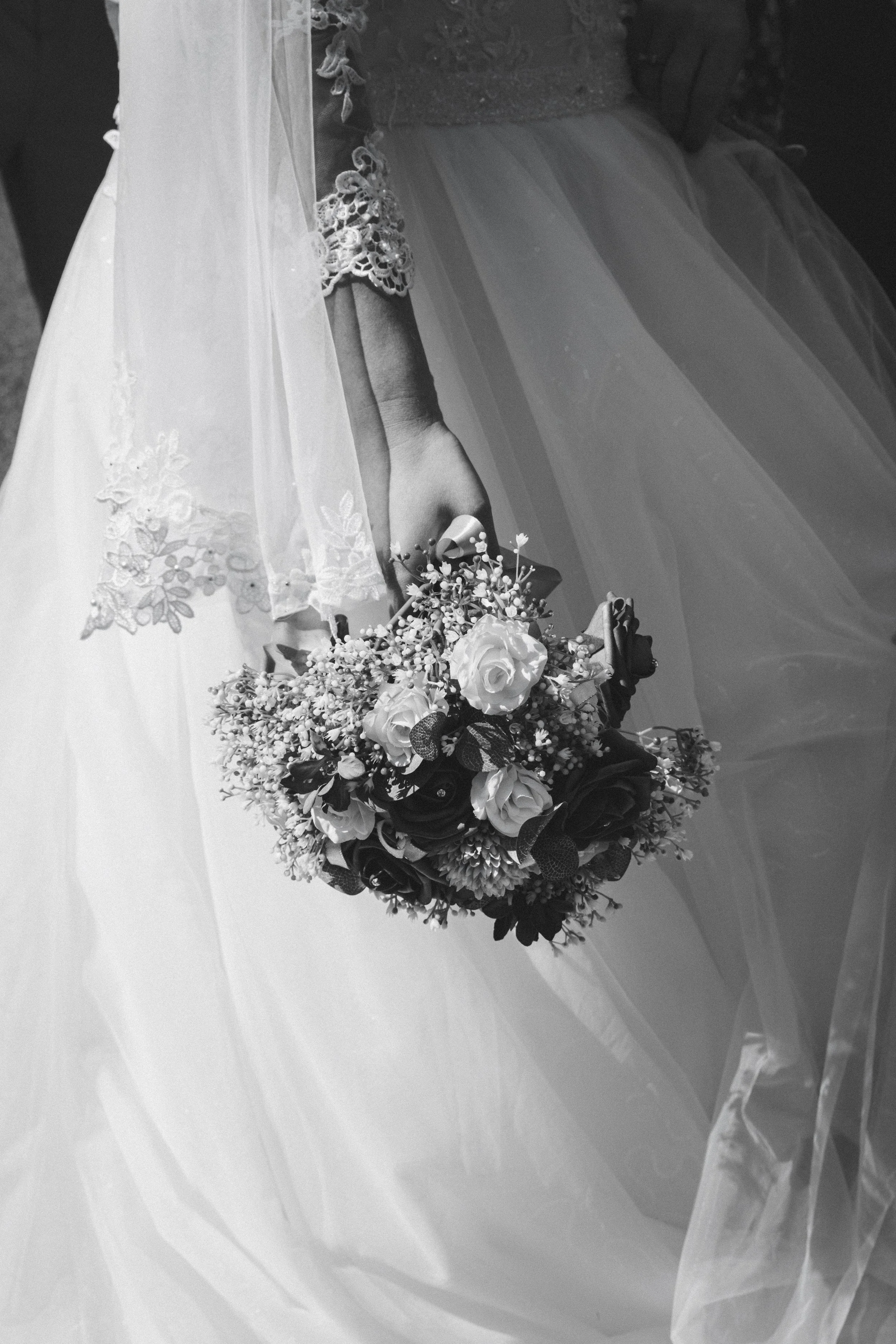 A bride holding a bouquet of roses and other flowers, wearing a wedding dress with lace details and a sheer veil, standing outdoors.