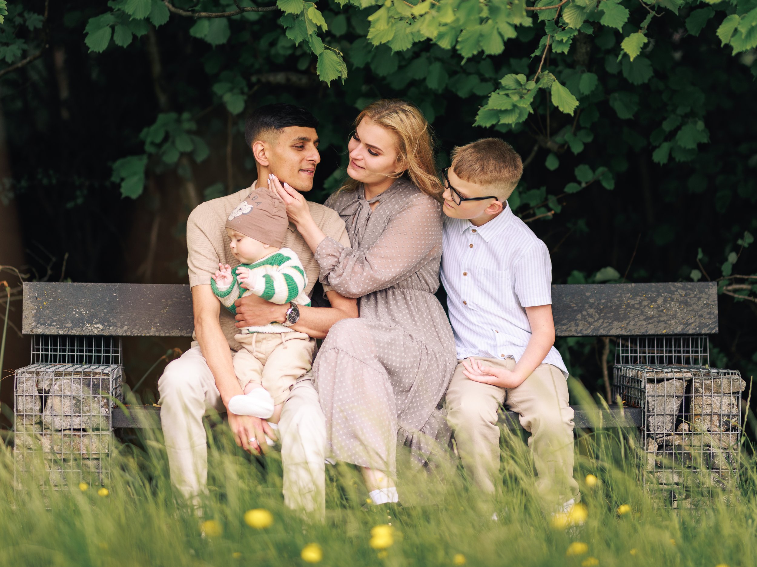 A family of five sitting on a wooden bench outdoors surrounded by greenery and yellow flowers. The father holds a sleeping baby, the mother gently touches the father's face, the older son sits beside the mother, and another young child is on the fath