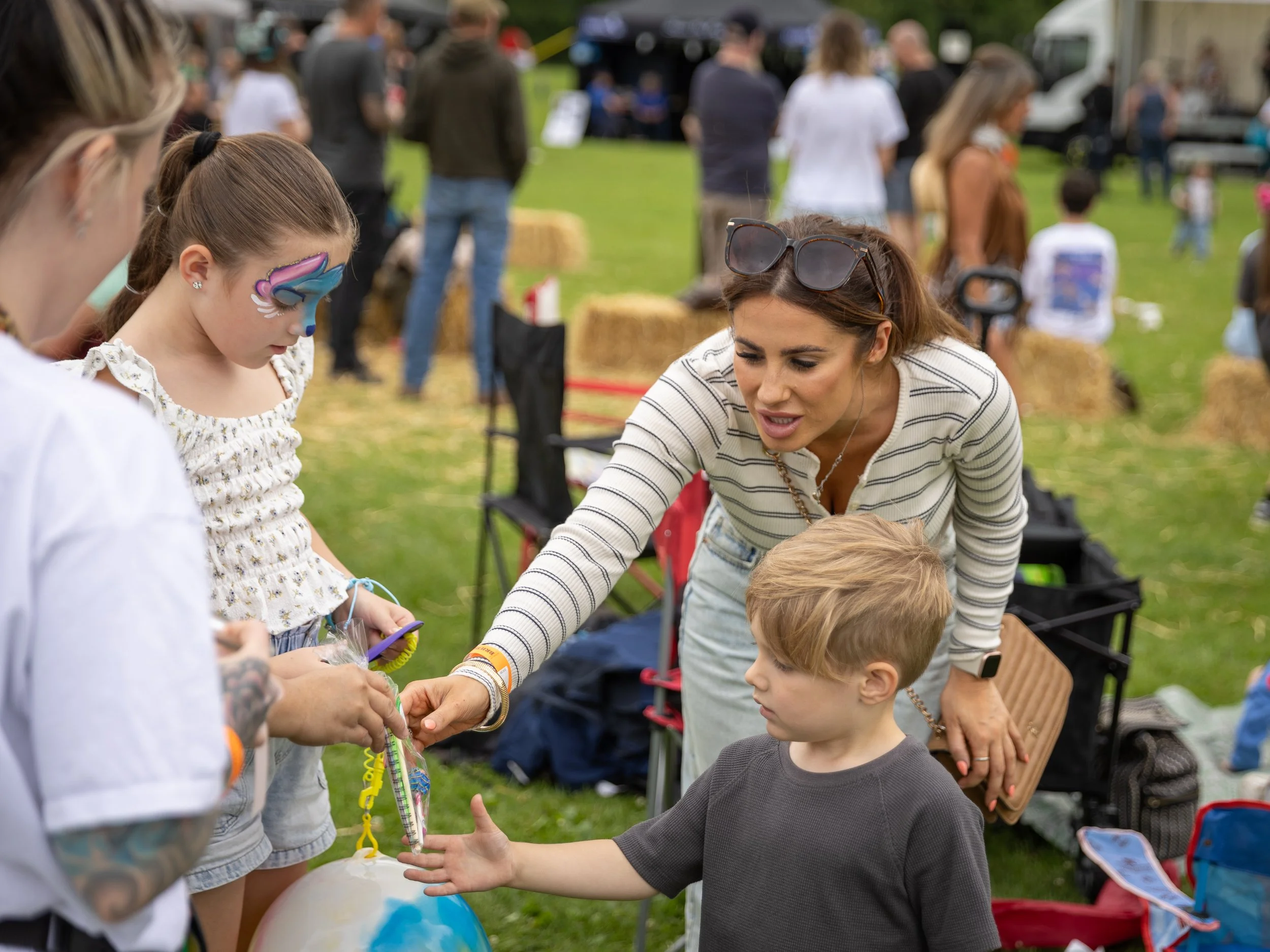 A woman with sunglasses on her head and a striped shirt is helping a young girl with face paint at an outdoor event. A boy is reaching out towards a decorated balloon. There are other people and hay bales in the background on a grassy field.