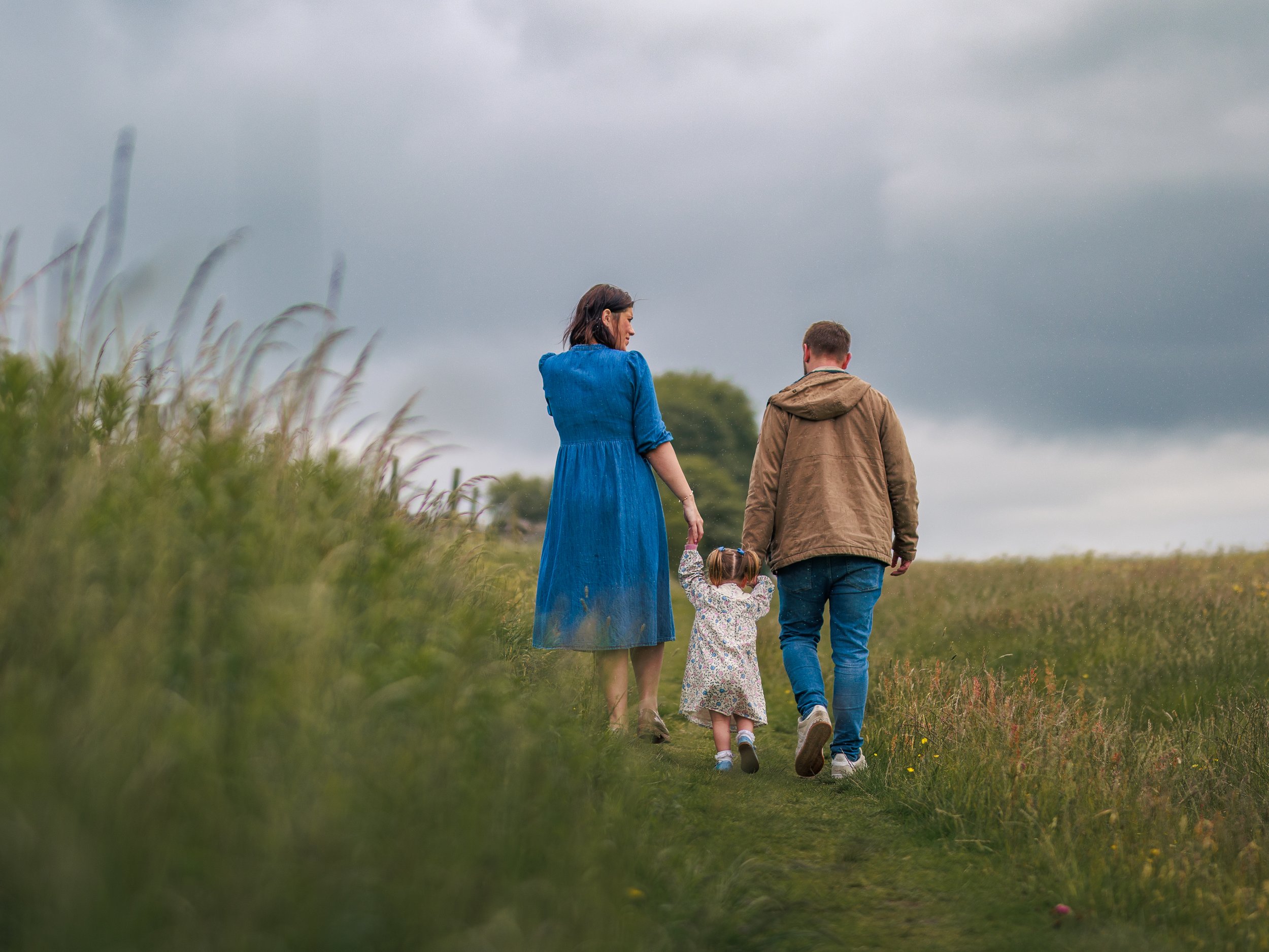 A family of three walking on a grassy trail outdoors under a cloudy sky. The mother is wearing a blue dress, the father a brown jacket and blue jeans, and their young daughter is holding hands with both.