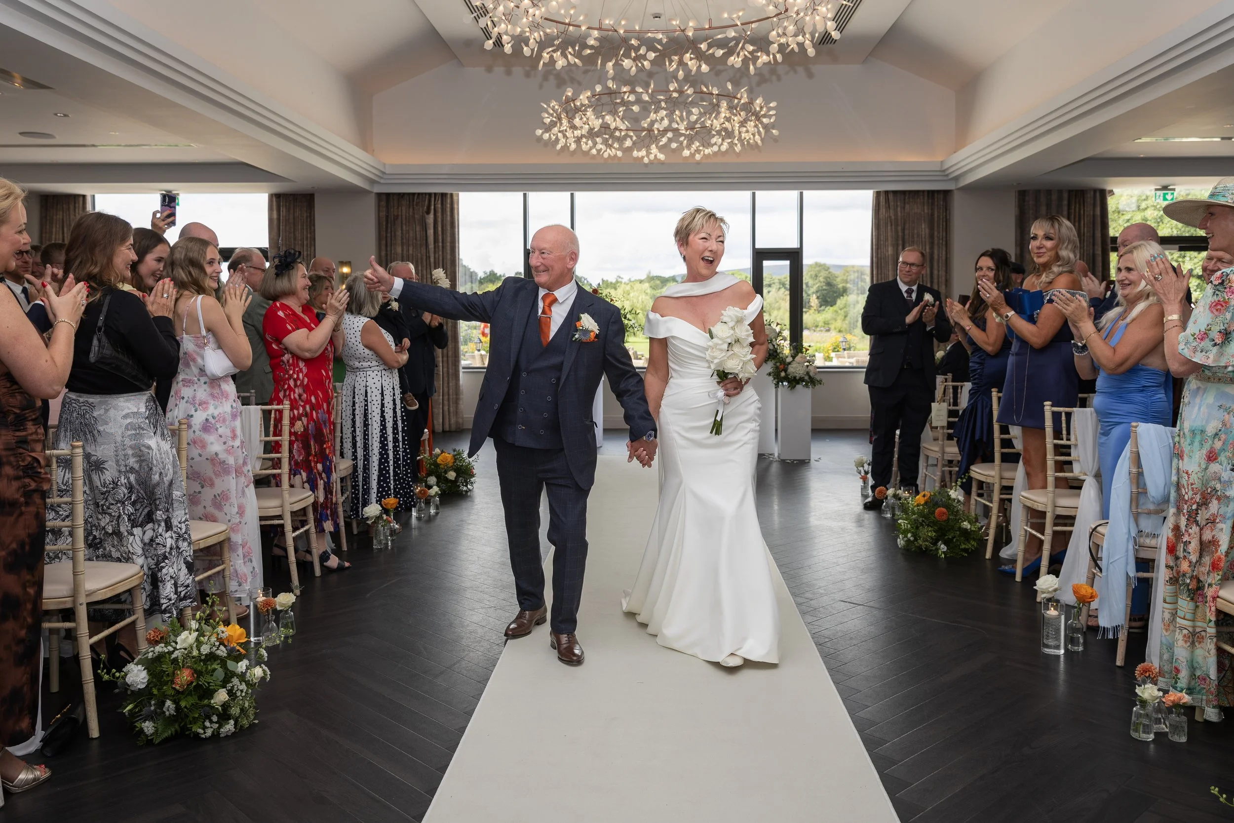 A bride and groom holding hands and walking down the aisle at a wedding, surrounded by smiling guests clapping and celebrating indoors with large windows and a modern ceiling light fixture.