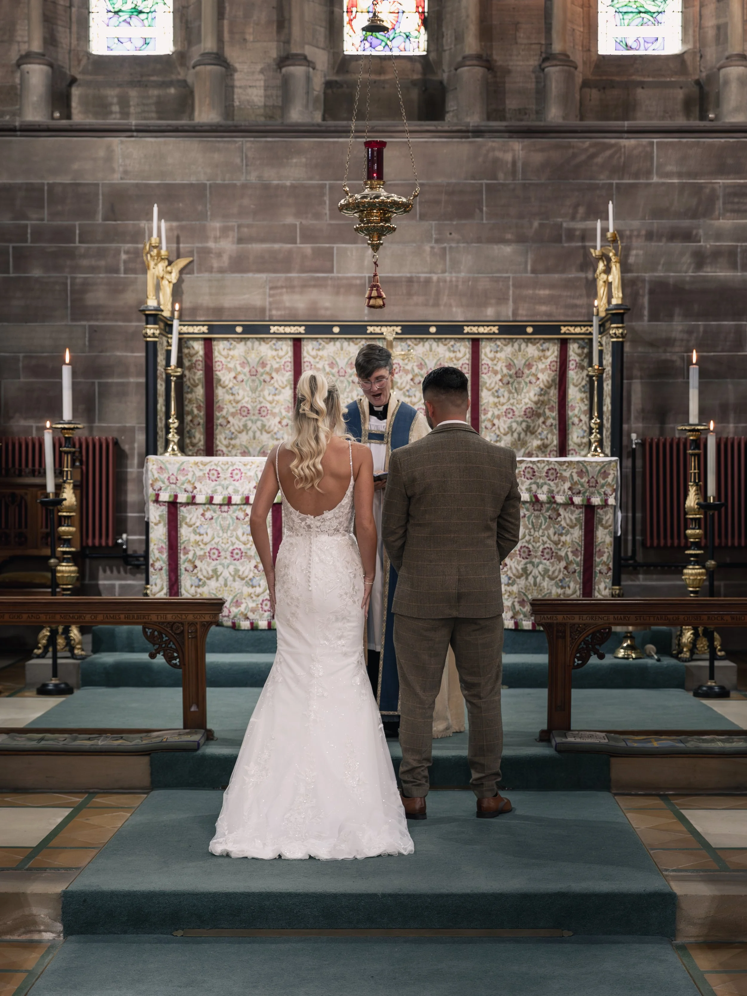 A couple getting married in a church with stained glass windows, standing at the altar with a priest. The bride is in a white wedding dress, and the groom is in a brown plaid suit. The altar has candles and religious decorations.