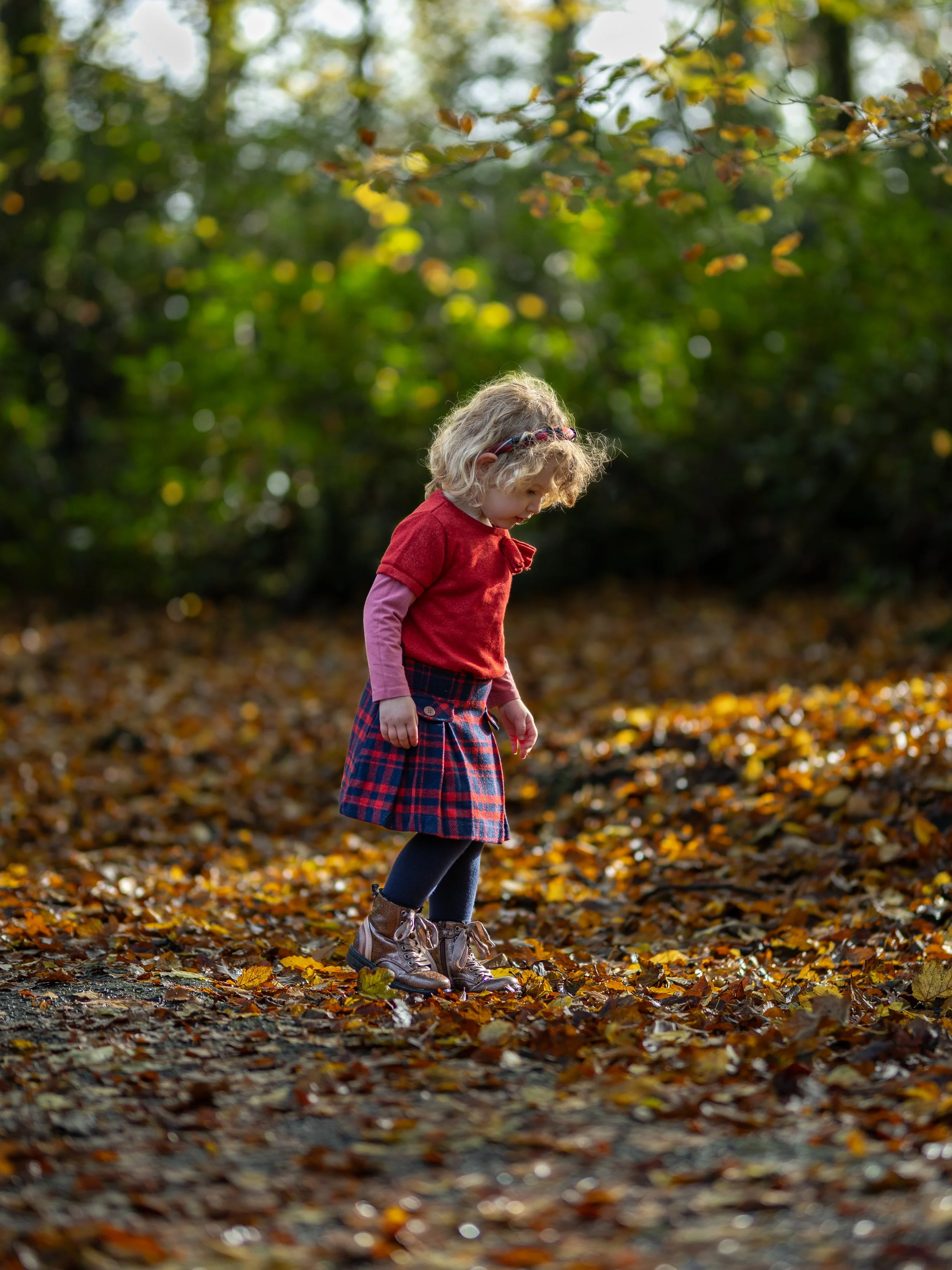 A young girl in a red sweater, pink long-sleeve shirt, plaid skirt, navy tights, and brown boots standing on a bed of fallen autumn leaves in a wooded area.