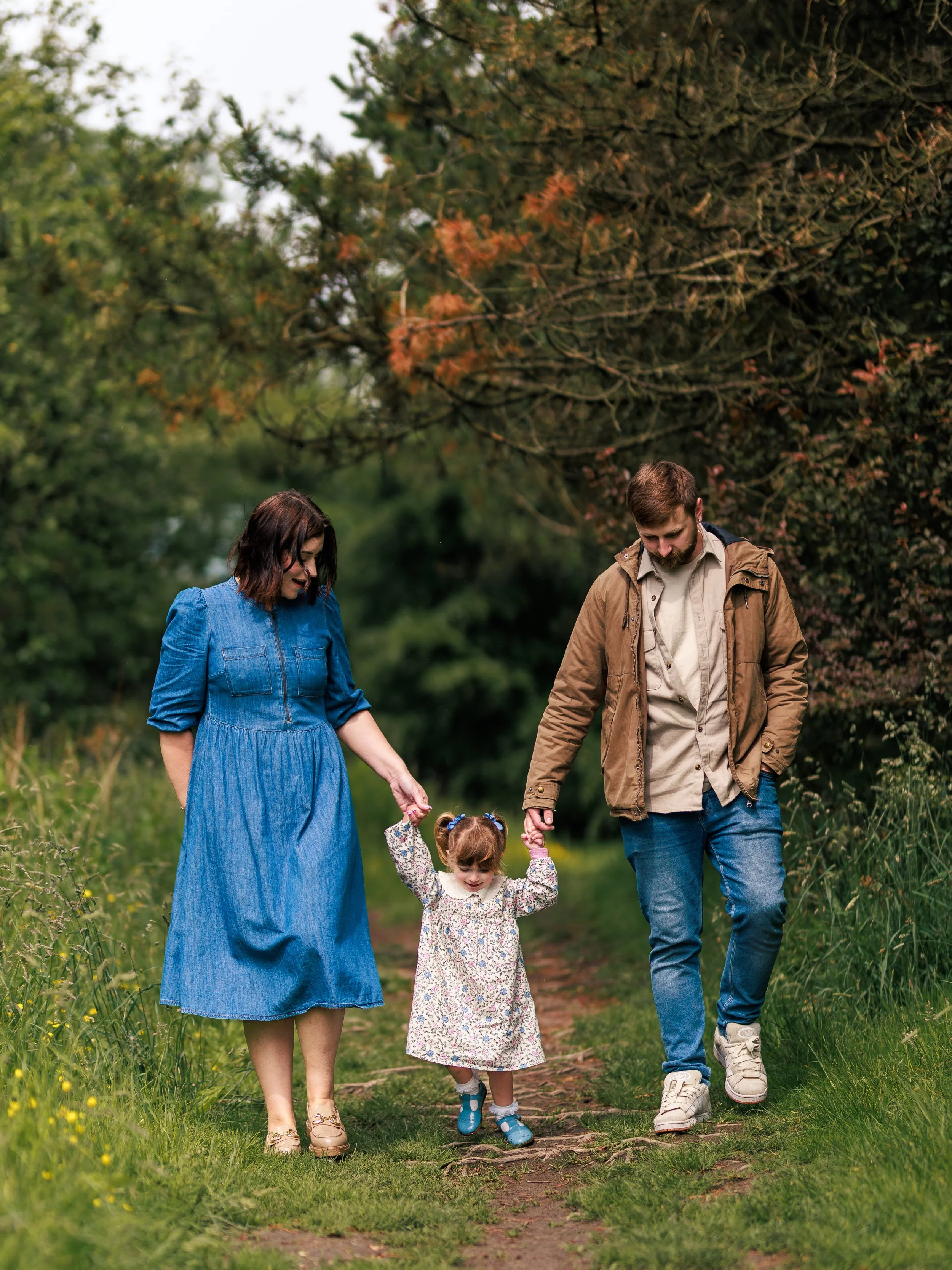 A family of three walking on a nature trail surrounded by trees and grass, holding hands and enjoying the outdoors.