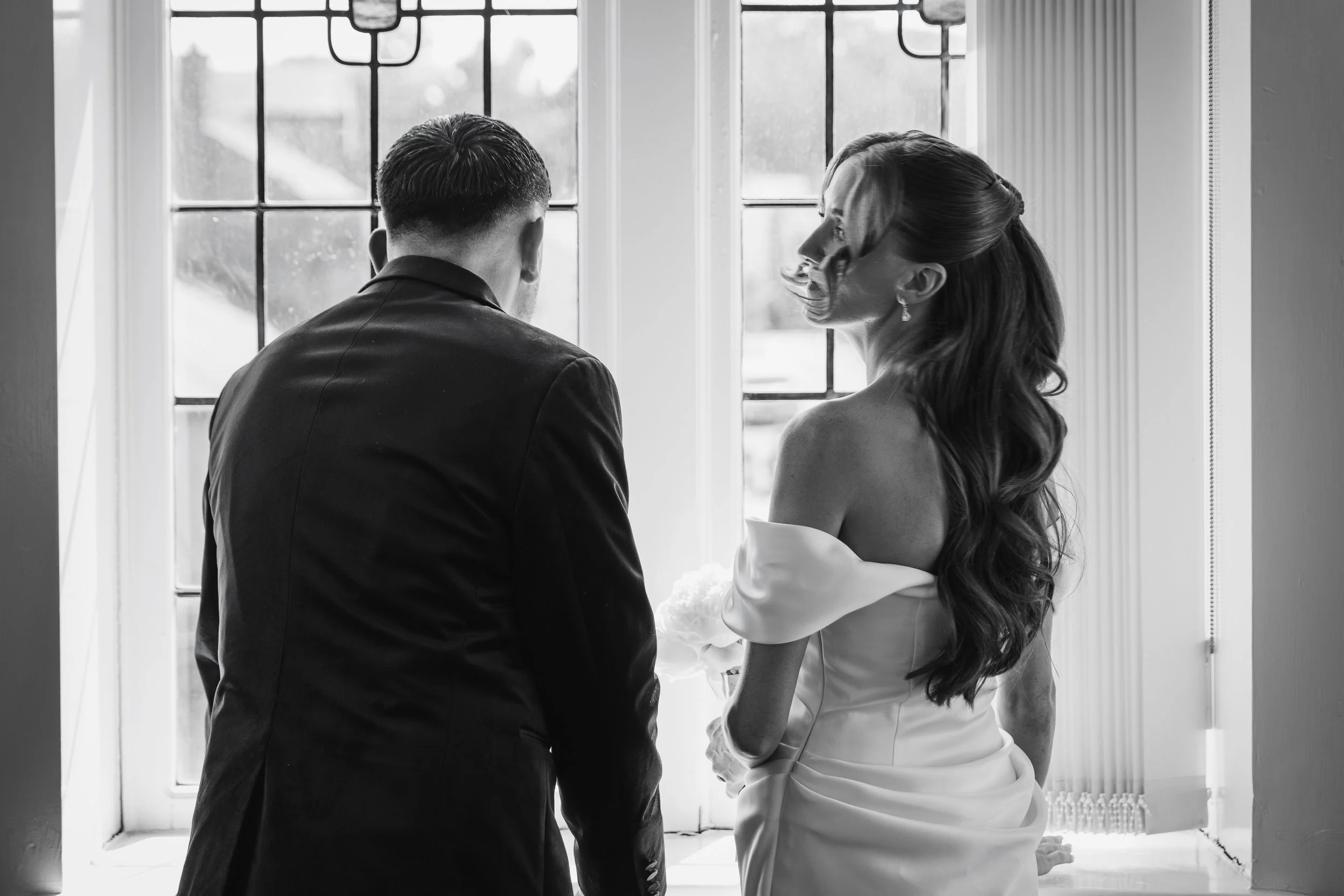 Black and white photo of a couple standing by a window, facing each other, dressed in wedding attire, sharing an intimate moment.