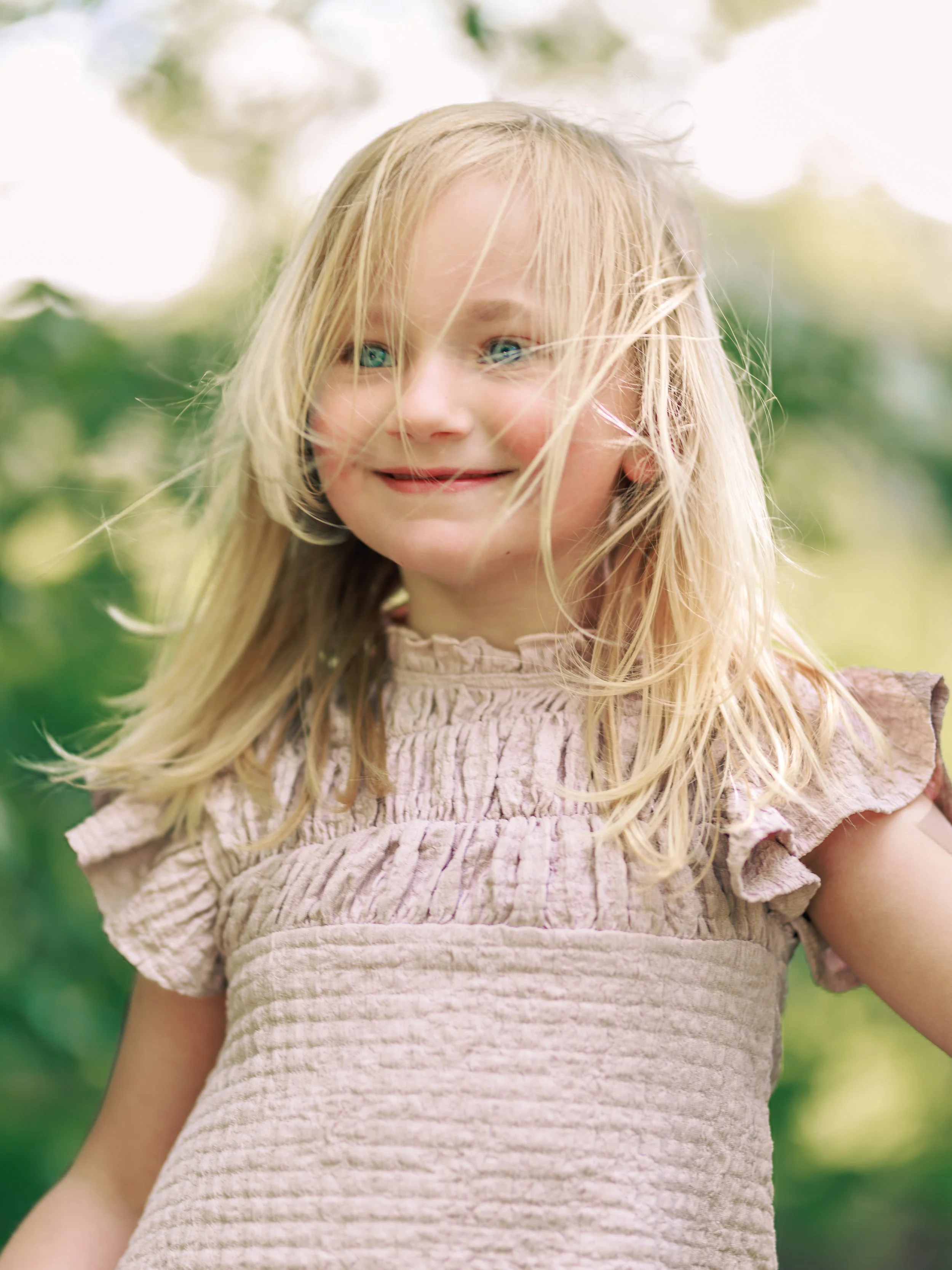 A young girl with blonde hair and blue eyes smiling outdoors on a sunny day.