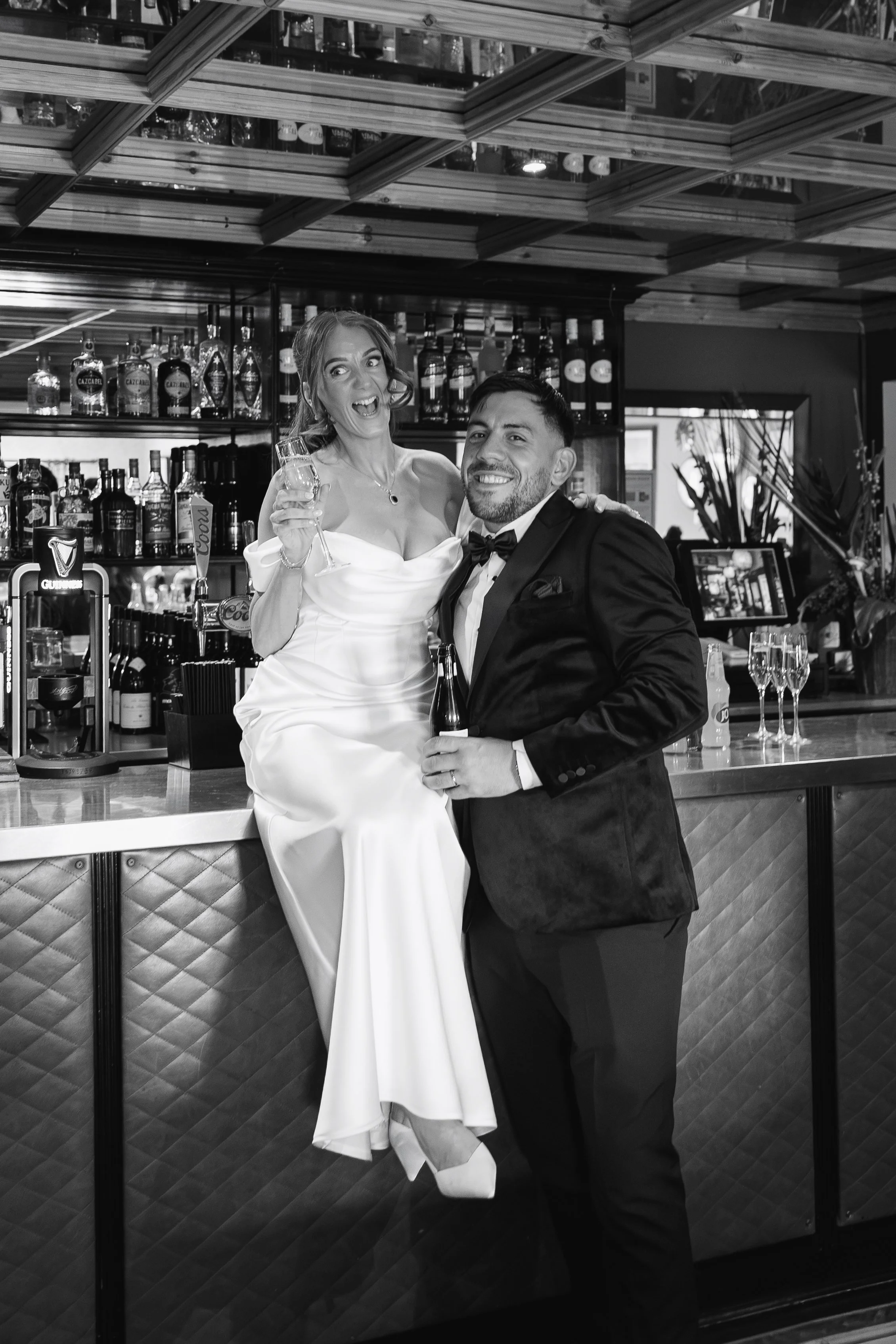 A woman in a wedding dress holding a glass of champagne, and a man in a tuxedo in a bar setting.