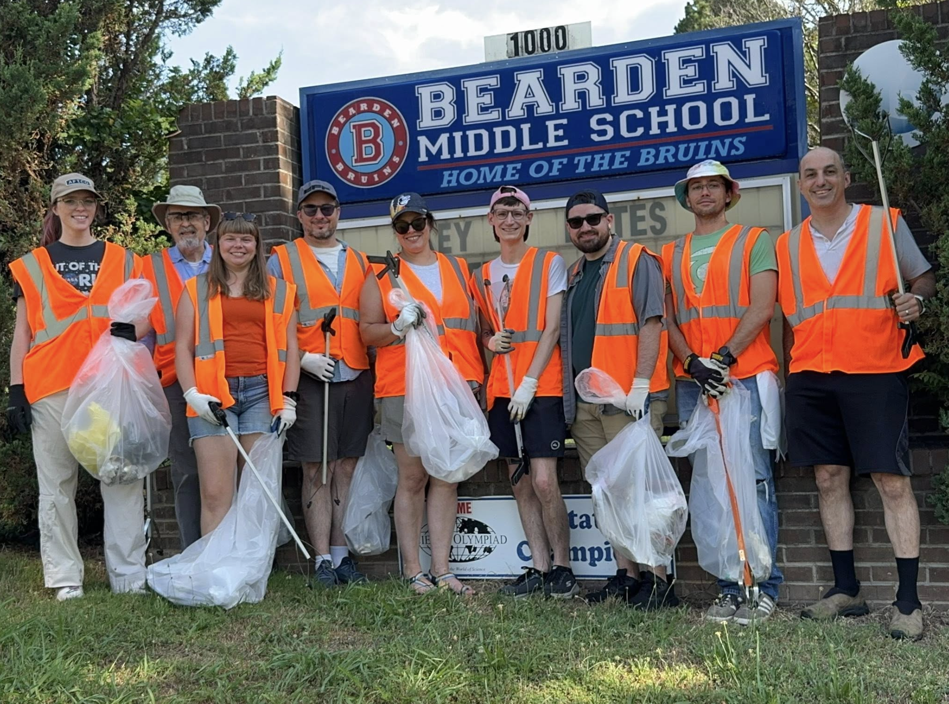 A group of ten people wearing orange safety vests and gloves standing in front of Bearden Middle School sign, participating in a community cleanup activity with trash bags and litter pickers.