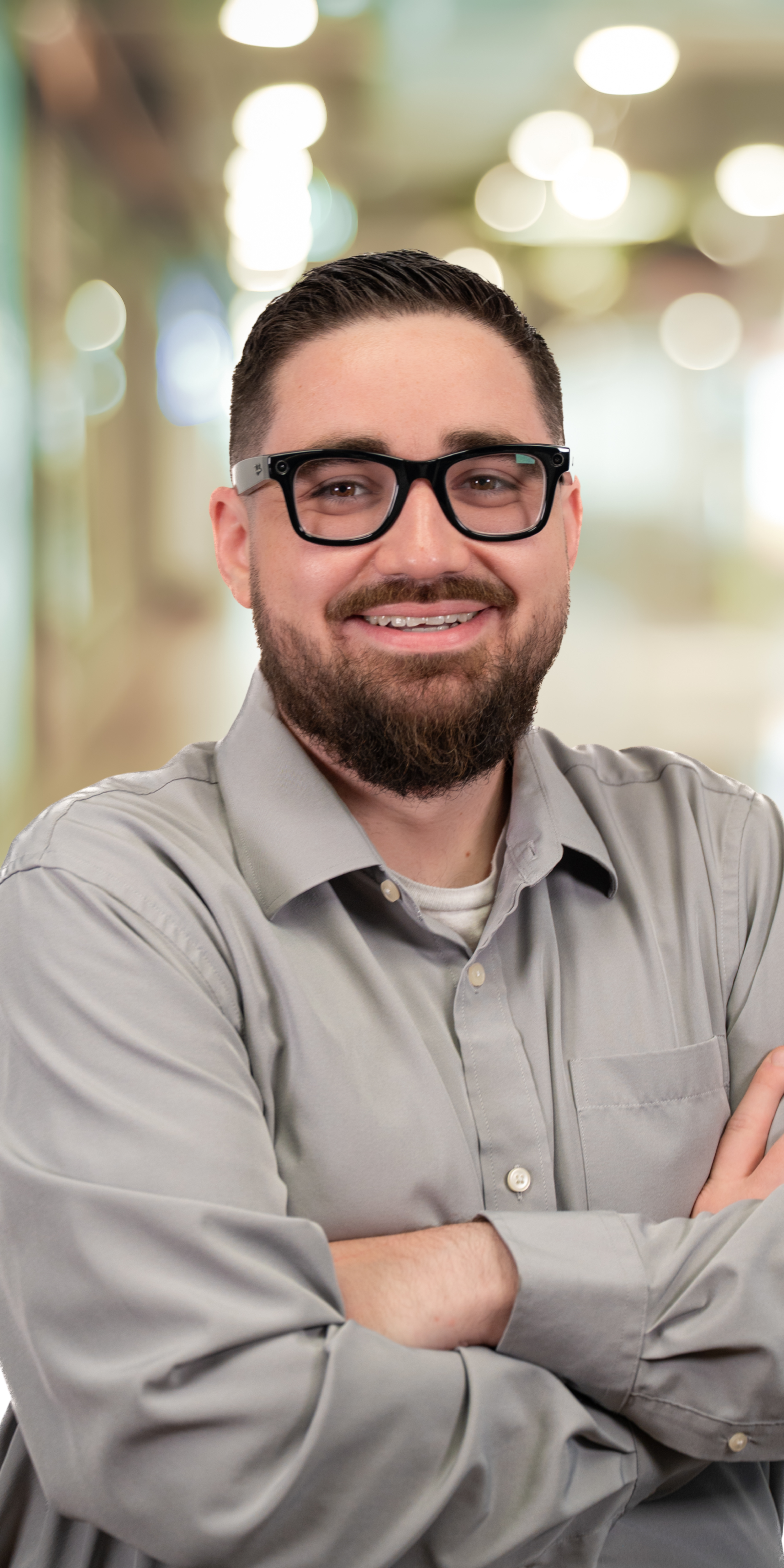 A young man with glasses and a beard smiling, standing with arms crossed in an indoor setting with blurred lights in the background.