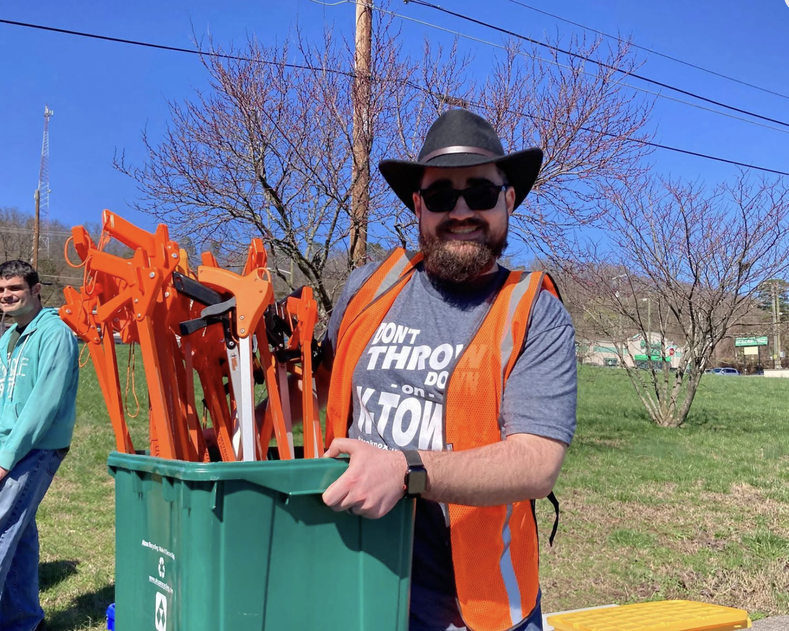 A smiling man wearing sunglasses, a wide-brimmed hat, an orange safety vest, and a gray T-shirt holding a green recycling bin filled with trash pick-up tools on a grassy park area.