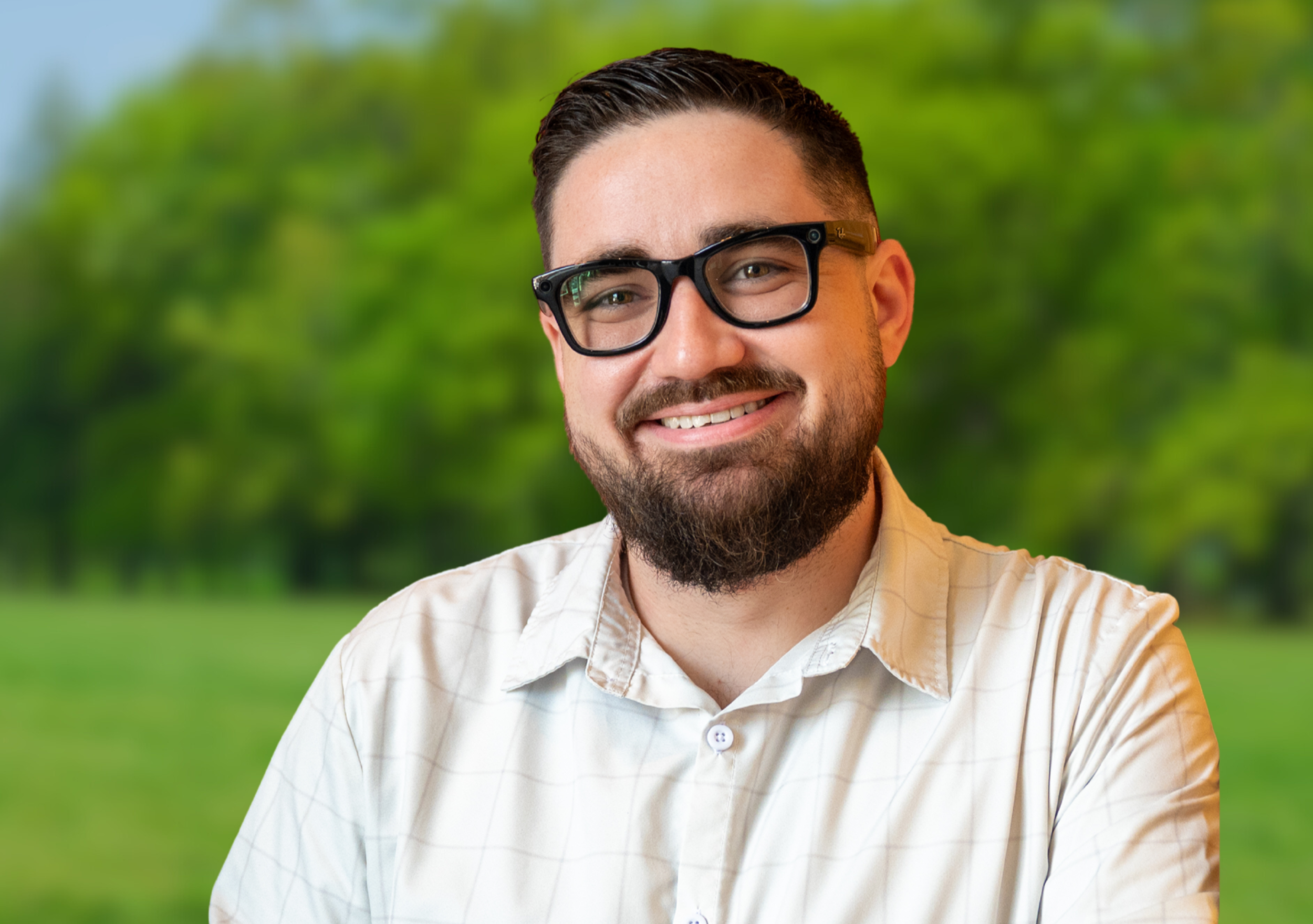 A smiling man with dark hair, a beard, and glasses, wearing a light-colored checkered shirt, standing outdoors with a blurred green background of trees and grass. This is Brandon Huckaby, candidate for Knox County Commission, District 3.