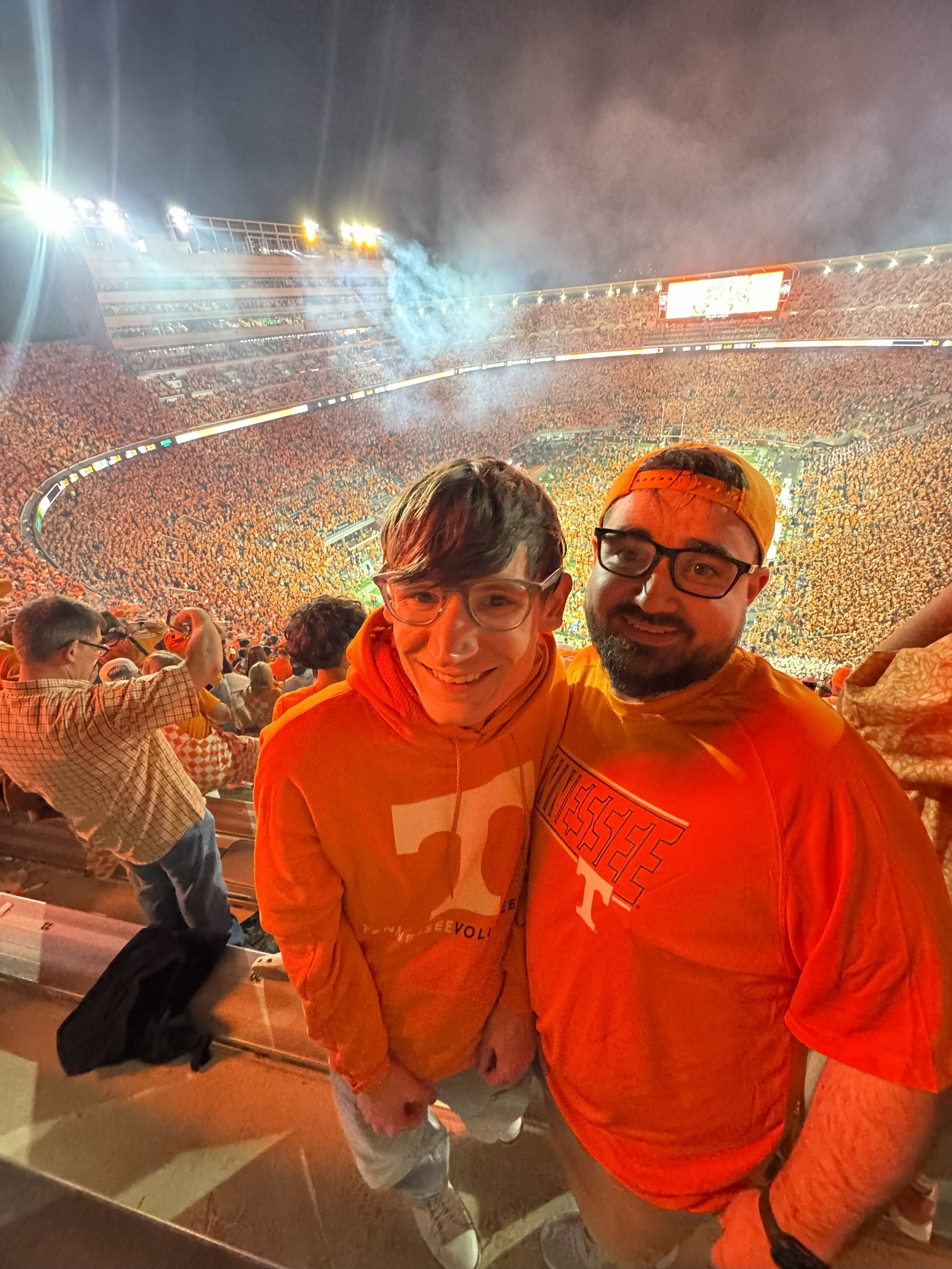Two smiling men at a stadium, wearing orange Tennessee Volunteers shirts, with a football game in the background.