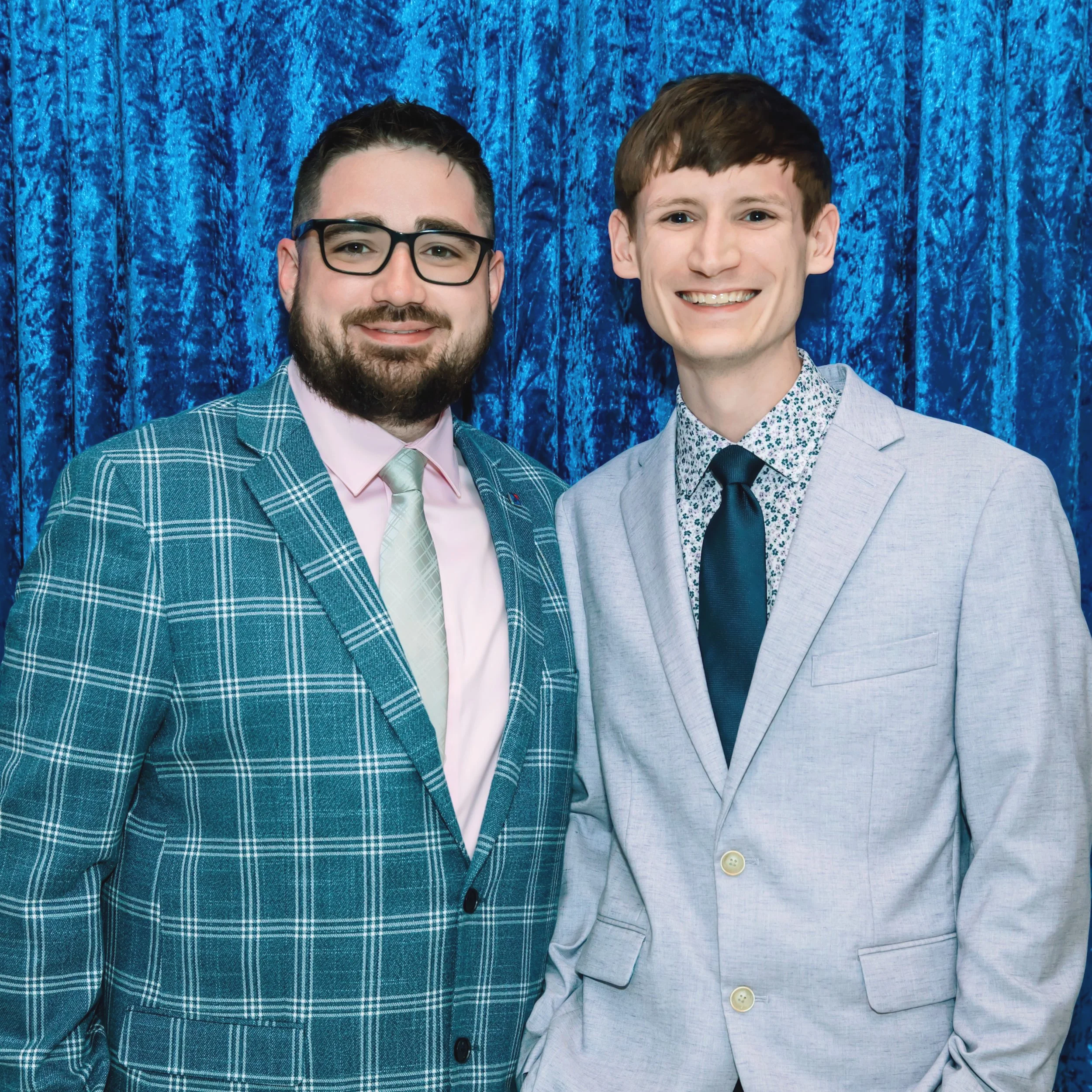 Two men in suits smiling in front of a blue curtain.