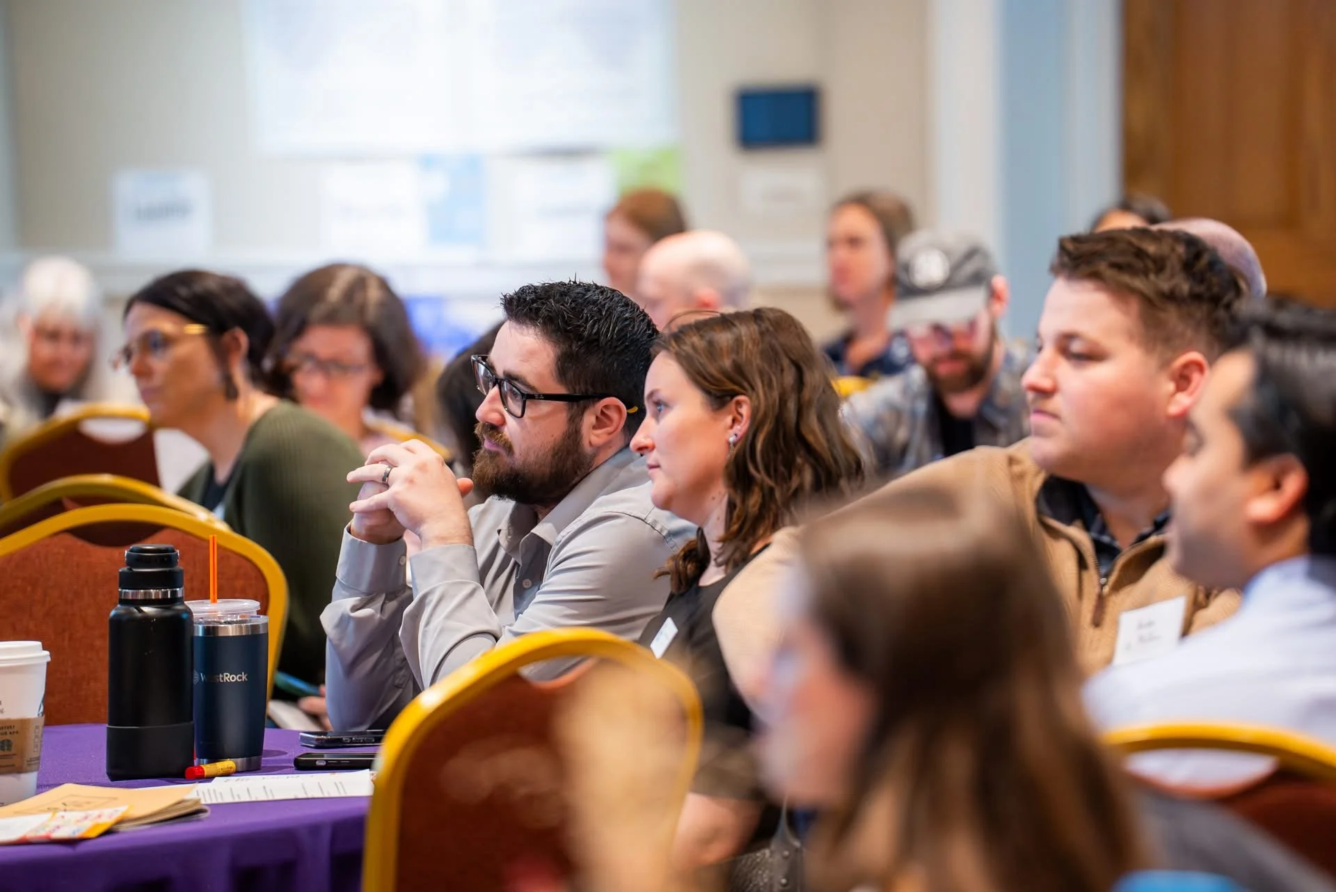 People attending conference or seminar, sitting at tables, listening attentively.