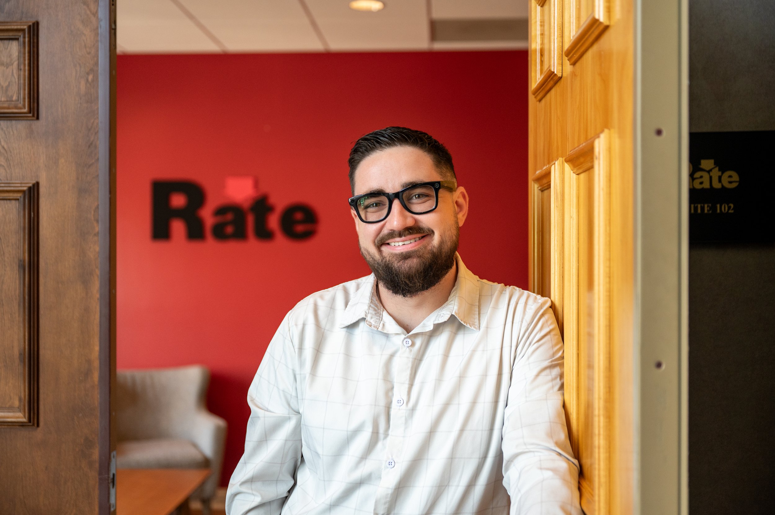 A man with glasses, beard, and short dark hair, smiling, standing in an office with red walls and wood accents. 'Rate' signage is visible in the background.