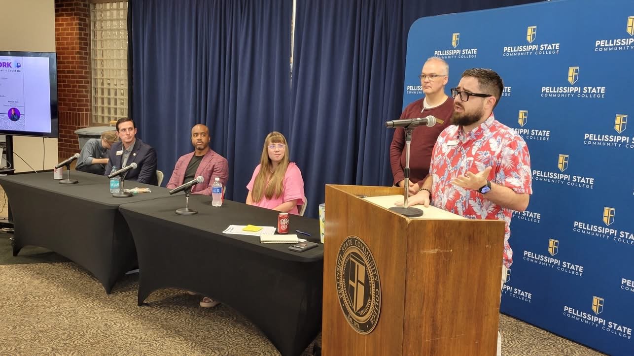 A man in a red tropical shirt speaking at a podium during a panel discussion at Pellissippi State Community College, with four other panel members seated behind him.