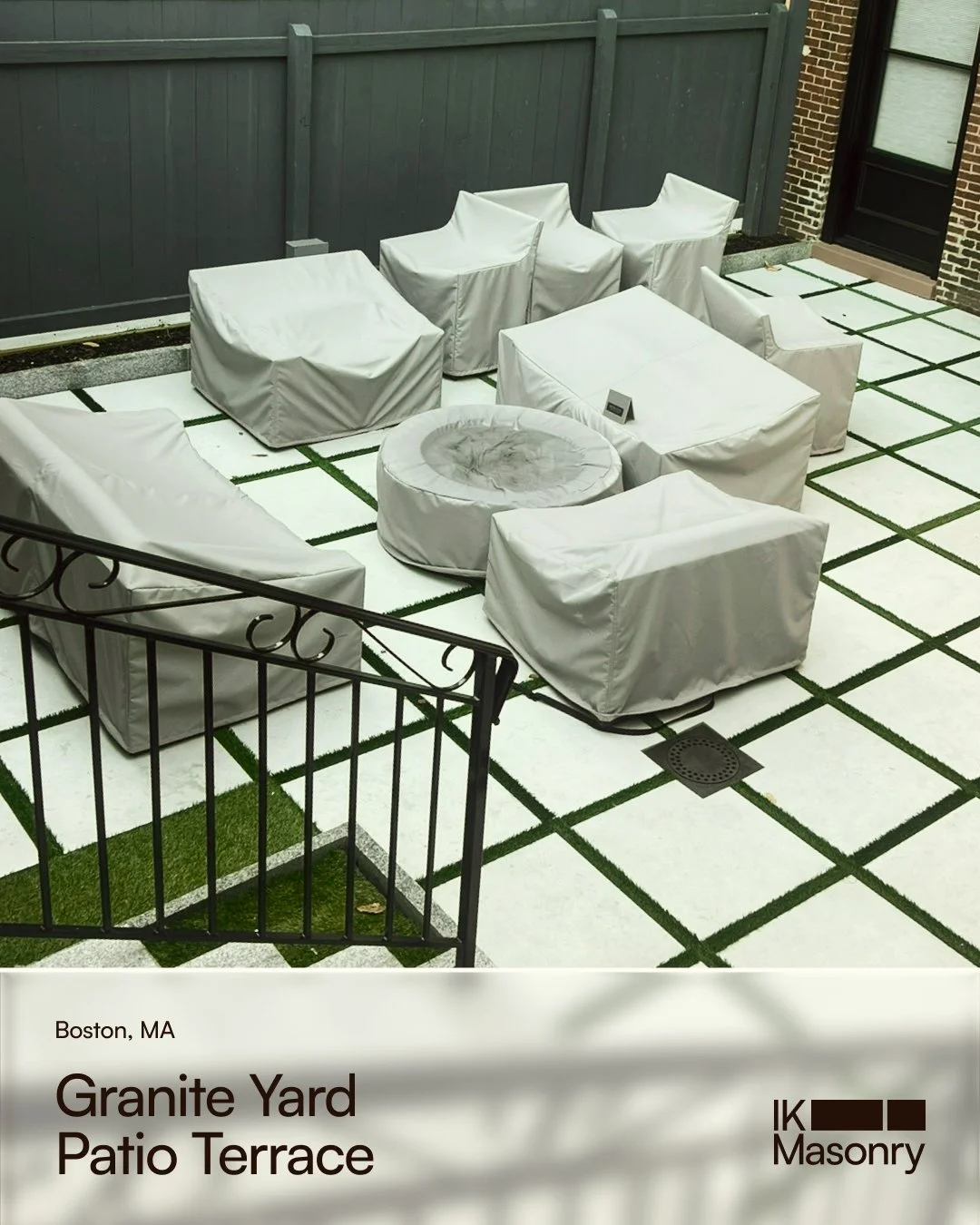 An outdoor patio with white covered furniture, including chairs, a round ottoman, and a coffee table, surrounded by a black metal fence, with a gravel and grass area and brick house exterior nearby.