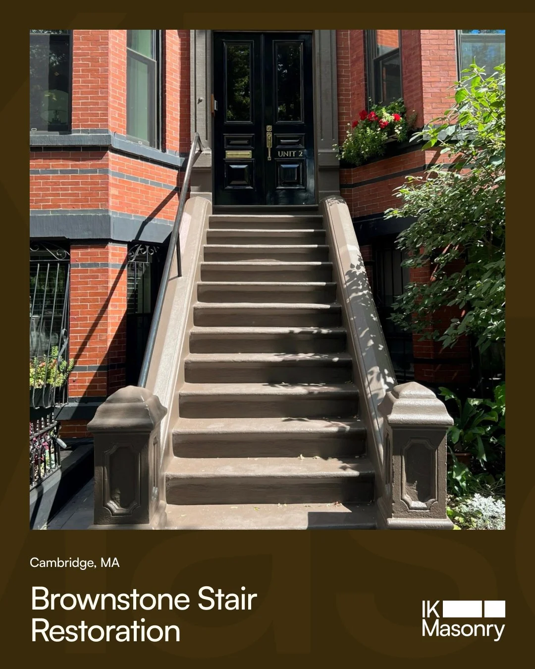Stone staircase leading to a black door with a gold mail slot and lock, outside brick building with a window box of flowers, greenery on the side, and metal handrails.