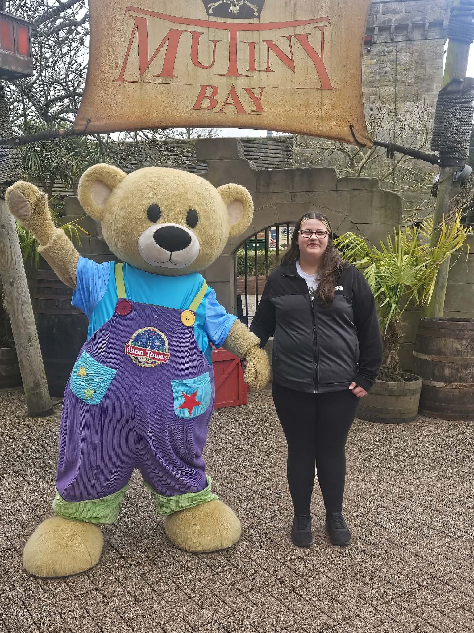 A person standing next to a large teddy bear mascot at Mummy Bay, Alton Towers resort. The mascot is wearing a blue shirt and purple overalls with colorful stars and a badge that says "Alton Towers". The person is wearing a black jacket and glasses, smiling and holding hands with the mascot. The background features a sign that reads "Mummy Bay" and some decorative plants.