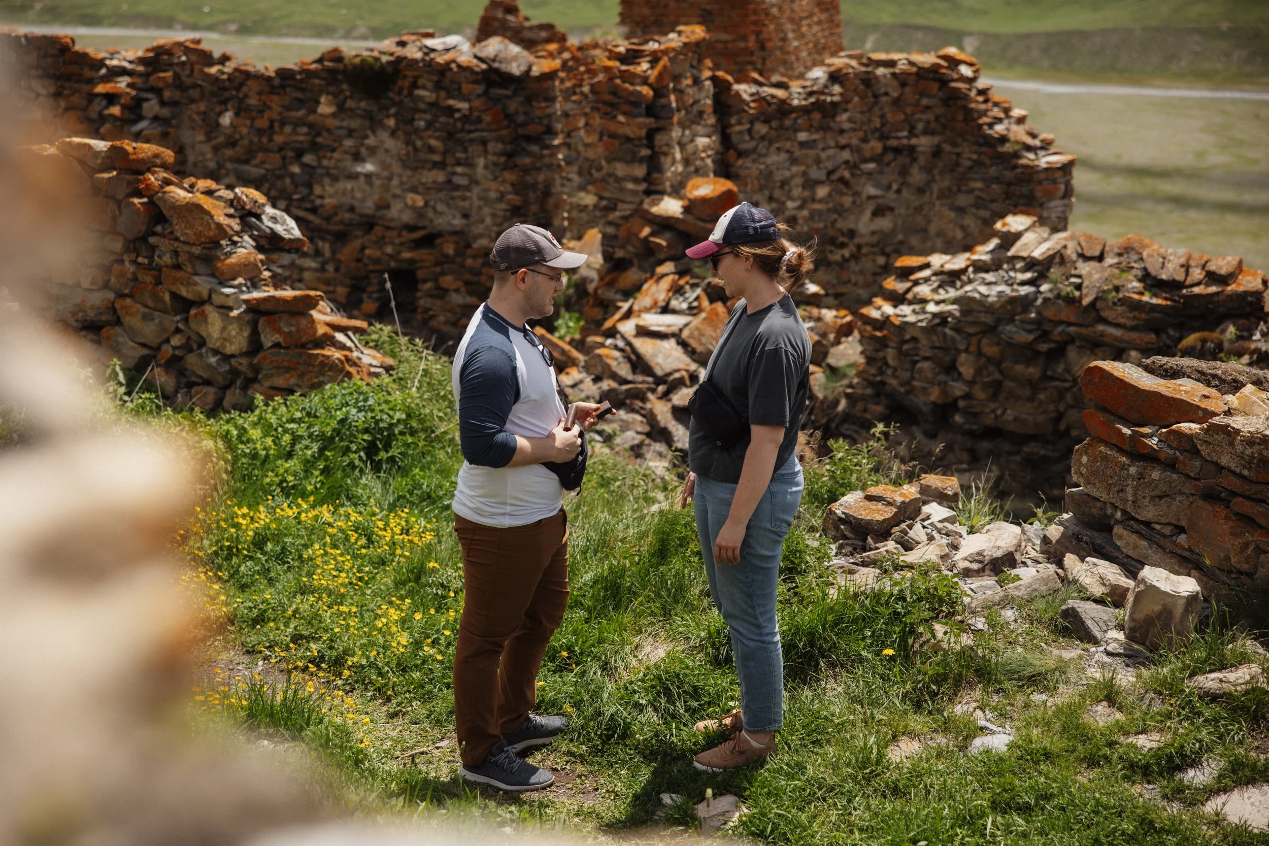 A man and woman stand outdoors near ancient stone ruins, engaging in conversation with each other.