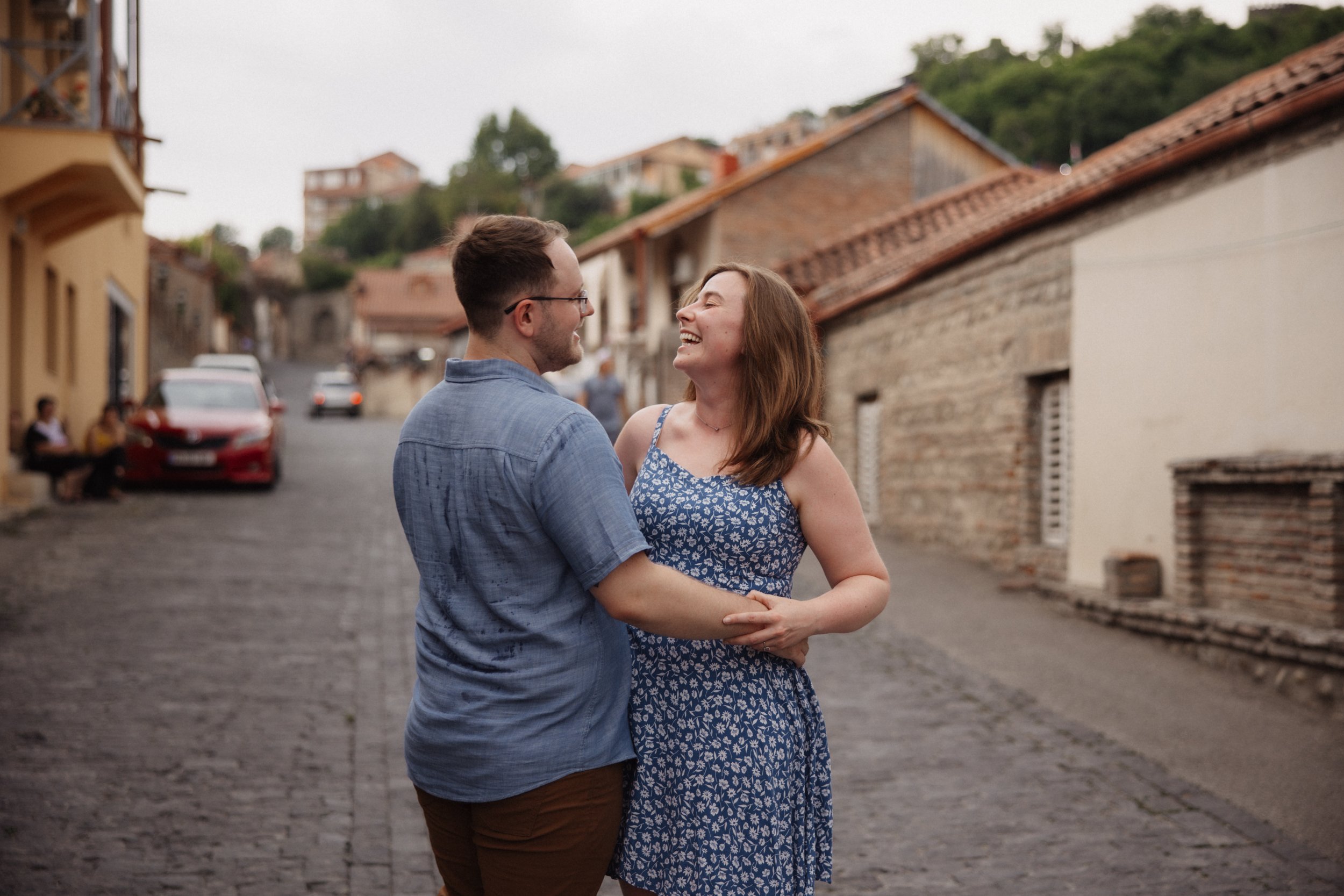 A young couple laughing and holding hands on a cobblestone street in a European town with colorful old buildings and cars parked along the side.