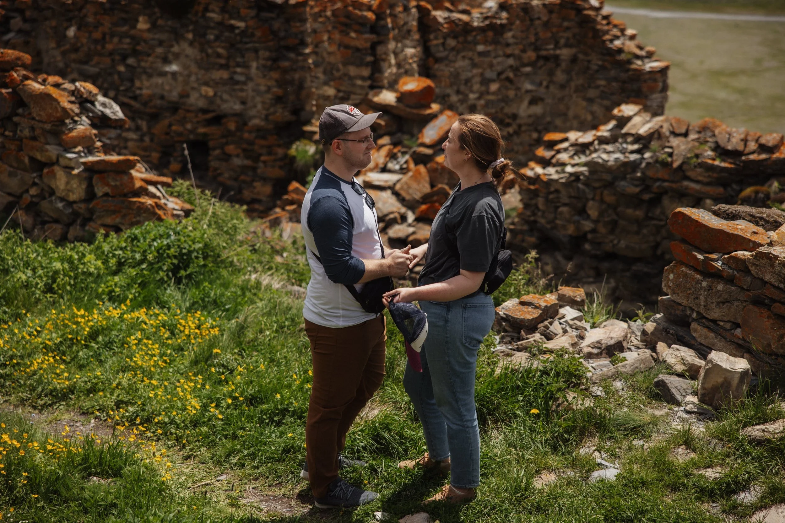 A man and woman stand close together outdoors, holding hands and gazing into each other's eyes near a ruin made of stacked stones, with green grass and yellow flowers around them.