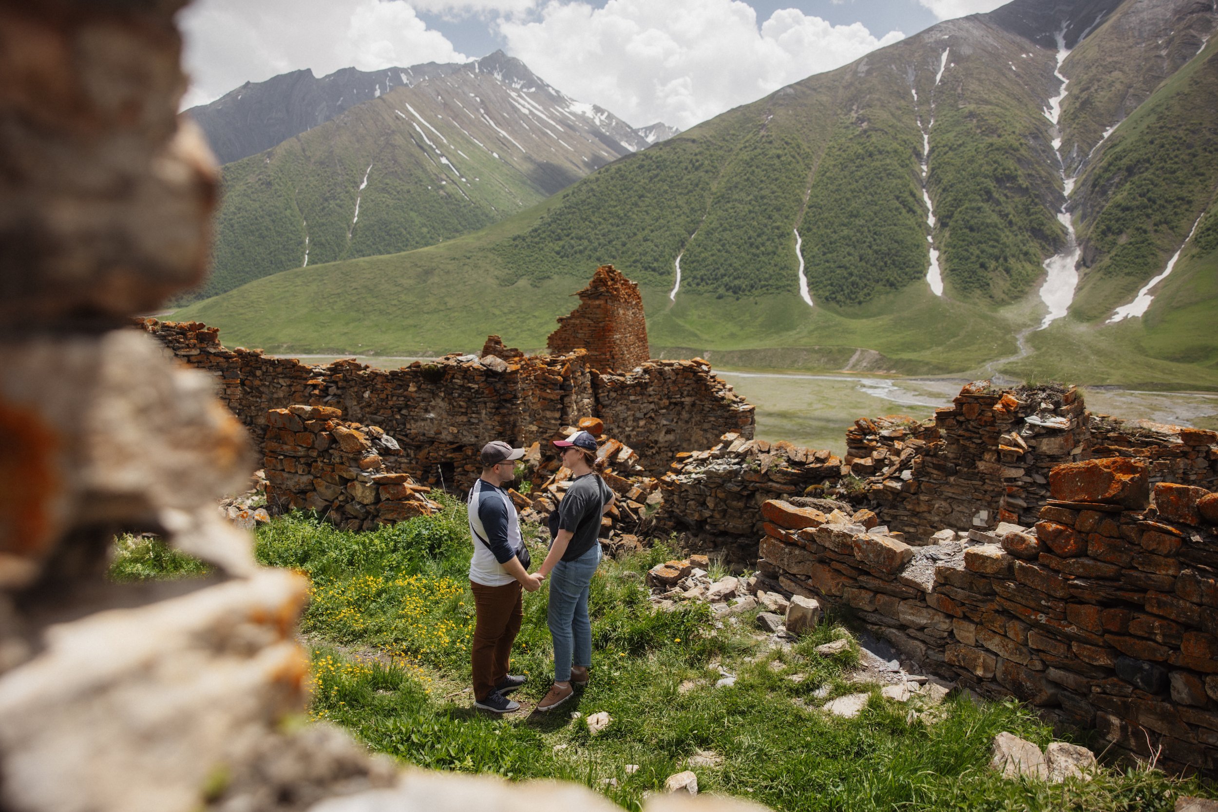 Two people holding hands and standing close together among ancient stone ruins, with green mountains and snow patches in the background.