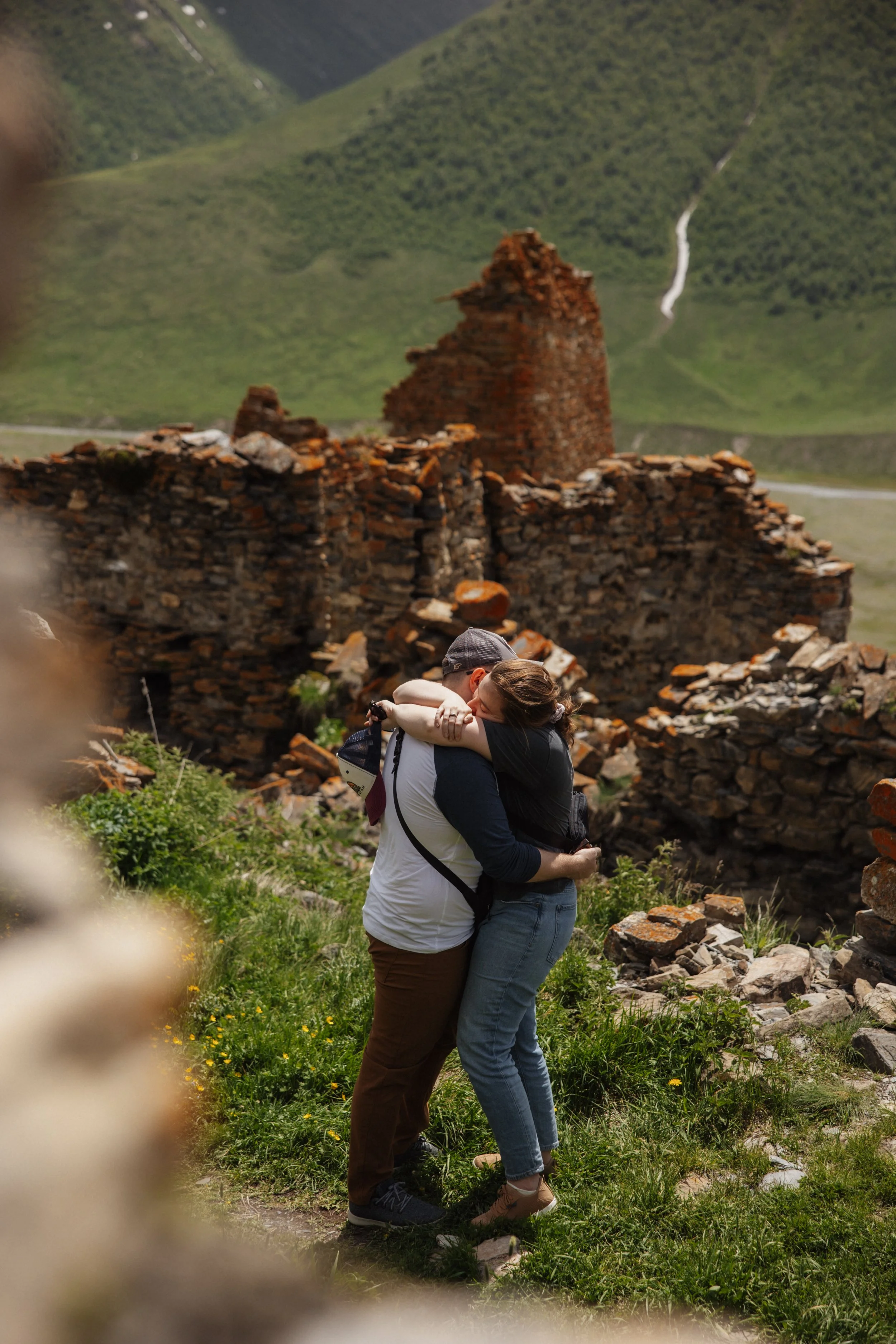 Two people hugging near an old stone ruin in a green, mountainous landscape.