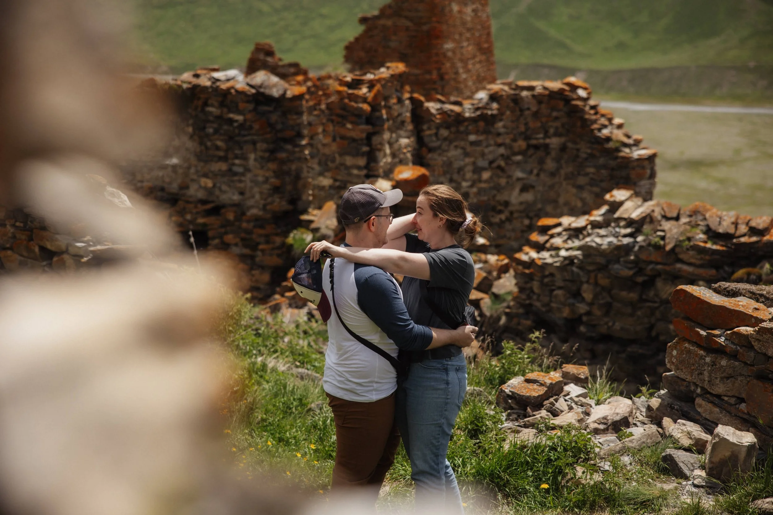A couple hugging outdoors near ruins with green hills in the background.