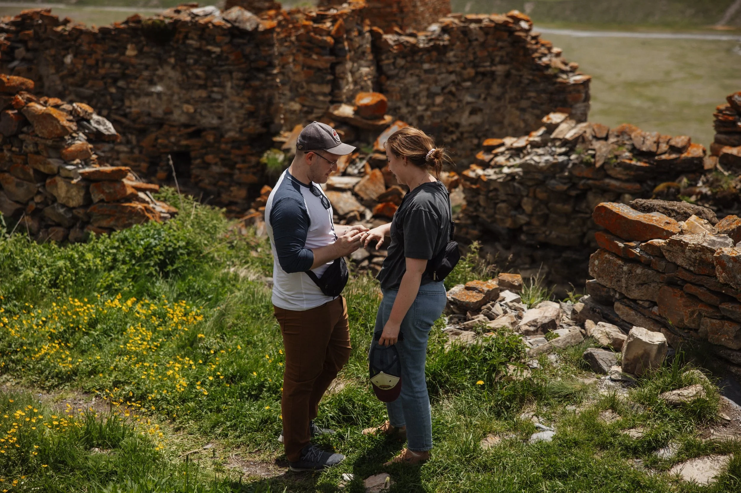 A man and woman standing outdoors among ruins, looking at a small object the man is holding, surrounded by green grass and yellow flowers.