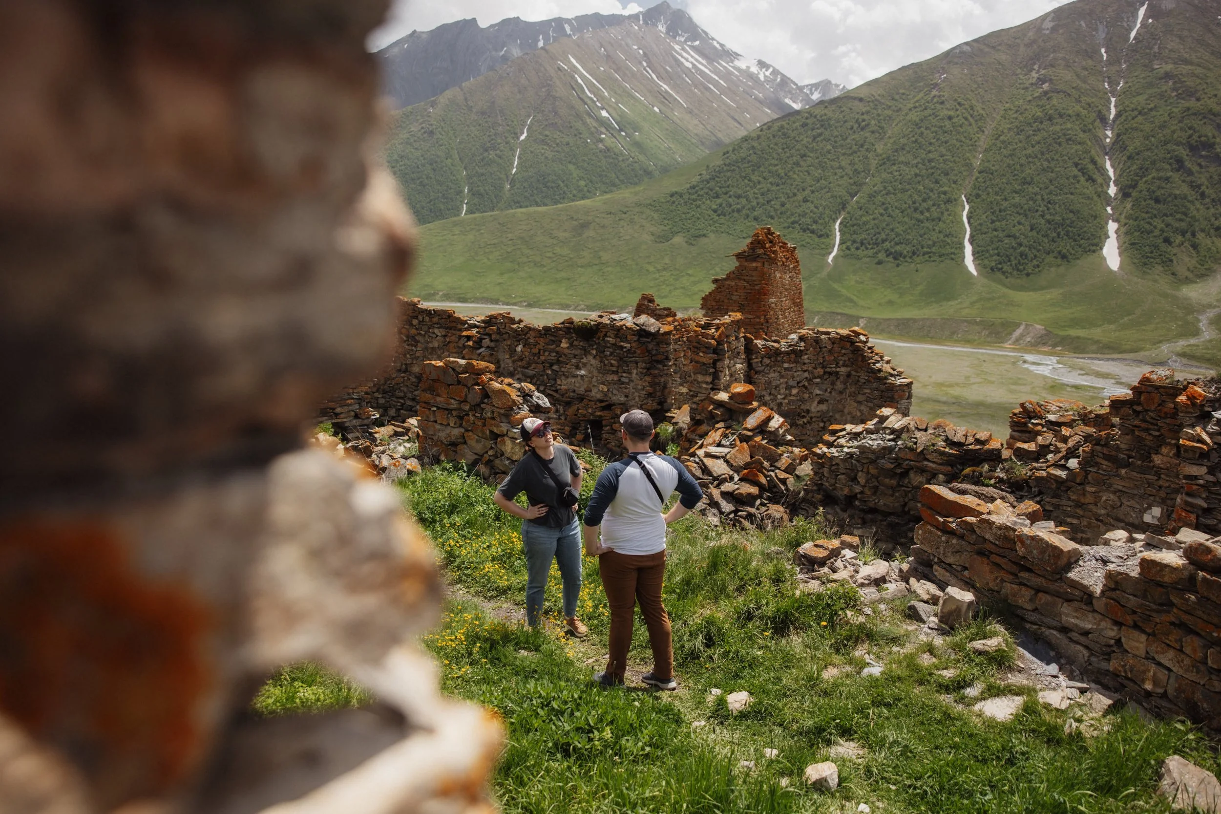 Two people talking among ancient ruins with old stone walls, surrounded by green grass, with mountain scenery in the background.