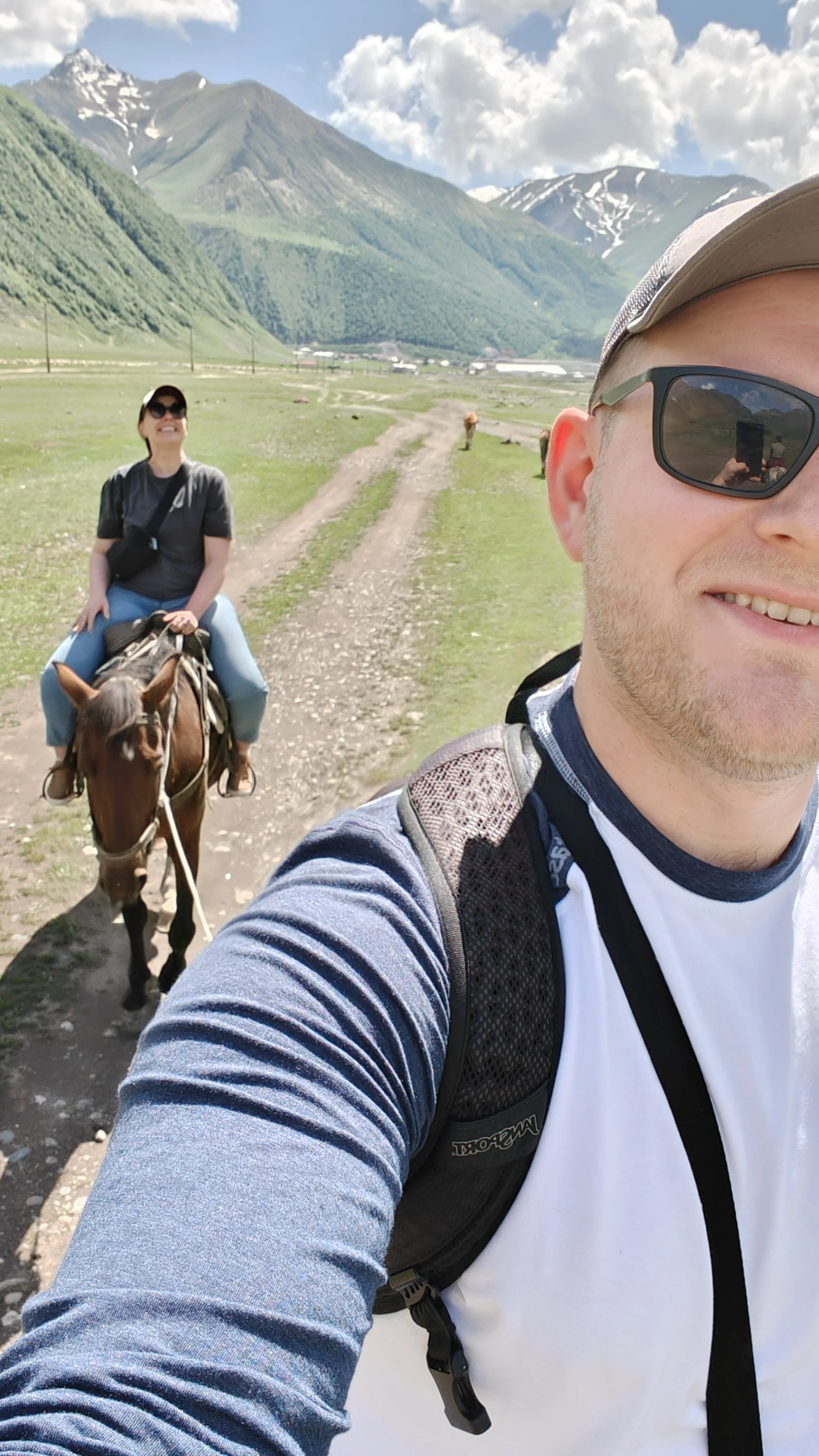 A man taking a selfie on a mountain trail with a woman riding a horse behind him. The woman is smiling, wearing sunglasses and a backpack, with mountains and a partly cloudy sky in the background.